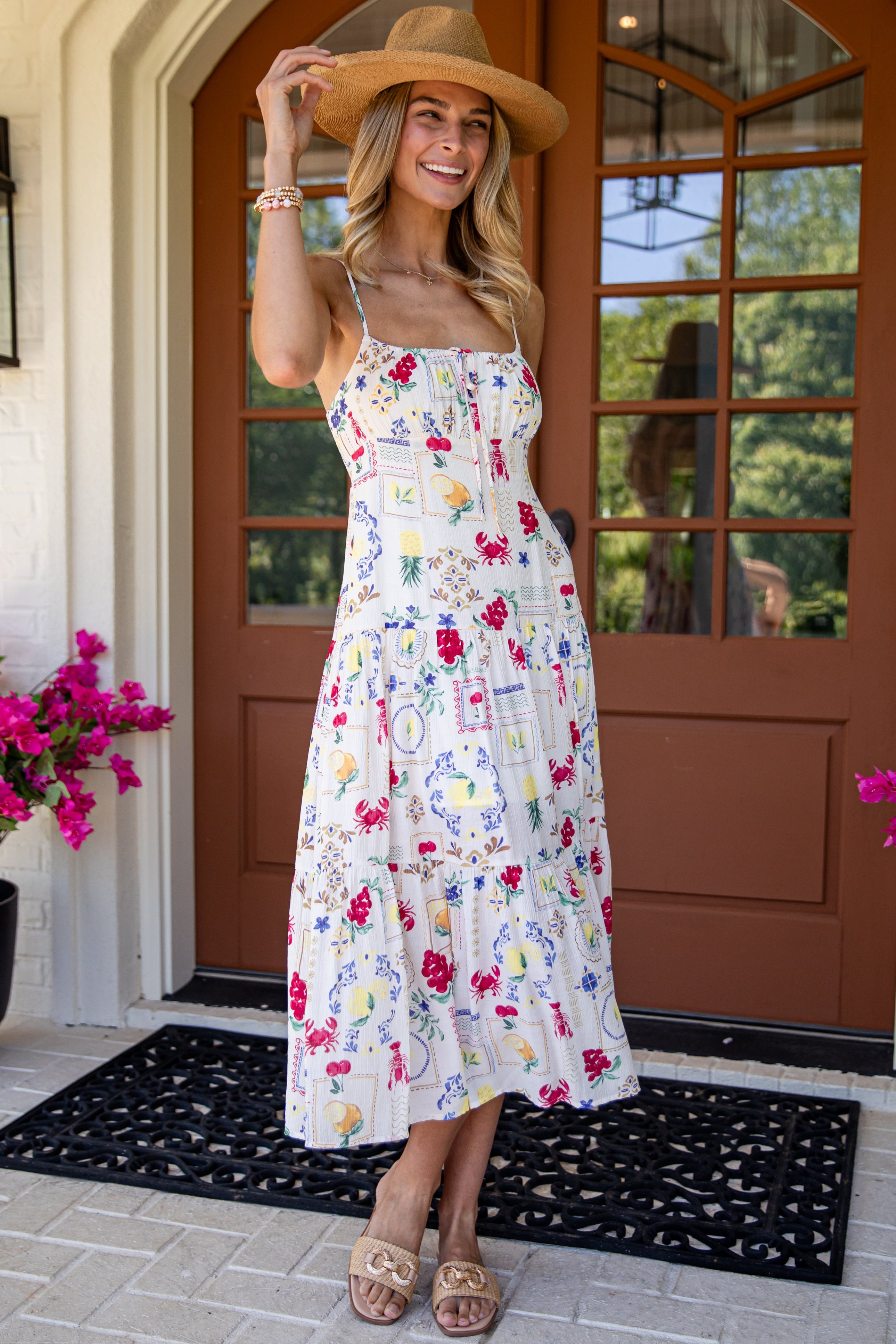 Woman in a floral dress and wide-brimmed hat standing in front of a house entrance.