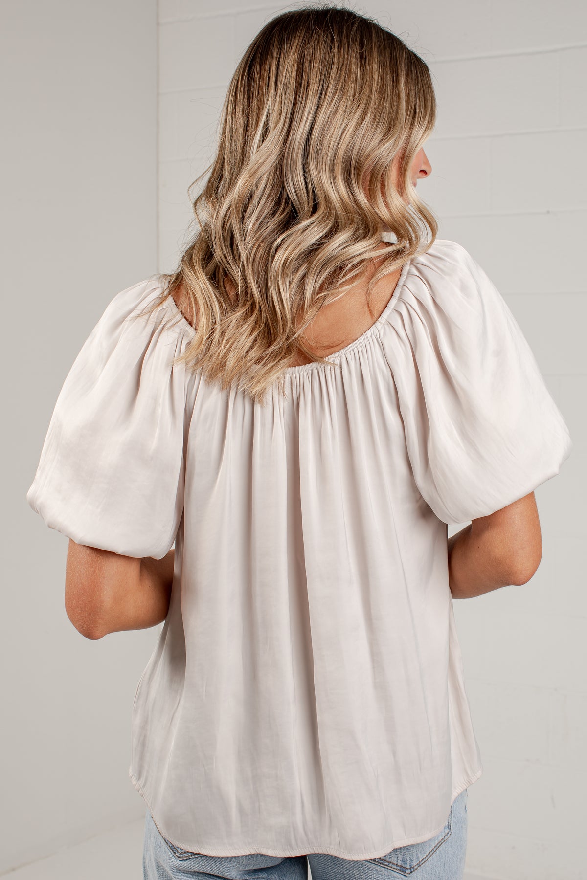 A woman viewed from behind wearing a light beige puff-sleeve top and light blue jeans, standing against a white paneled wall.