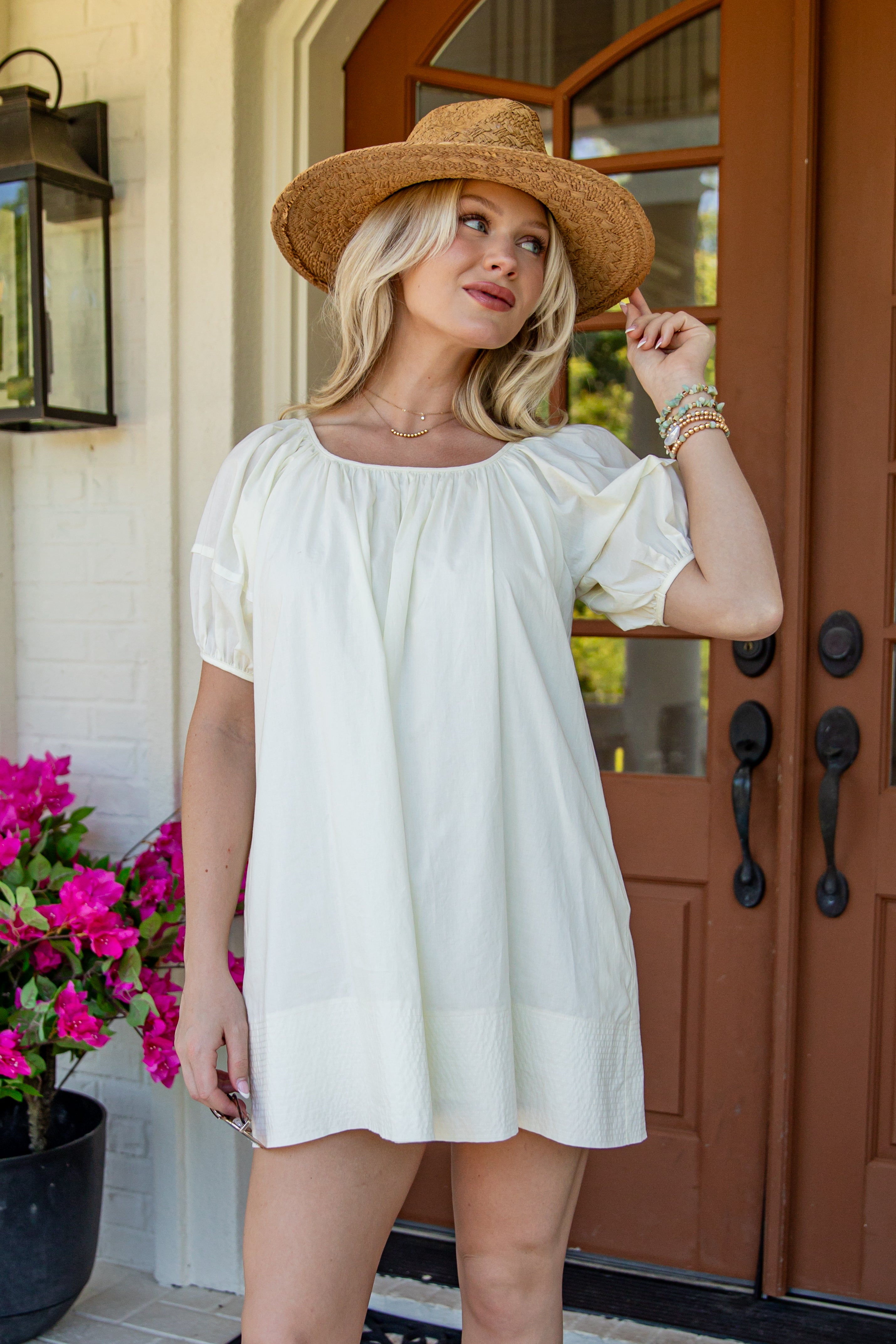 A woman wearing a cream-colored short-sleeve puff-sleeve tunic dress and a straw hat, standing on a front porch in front of brown wooden doors.