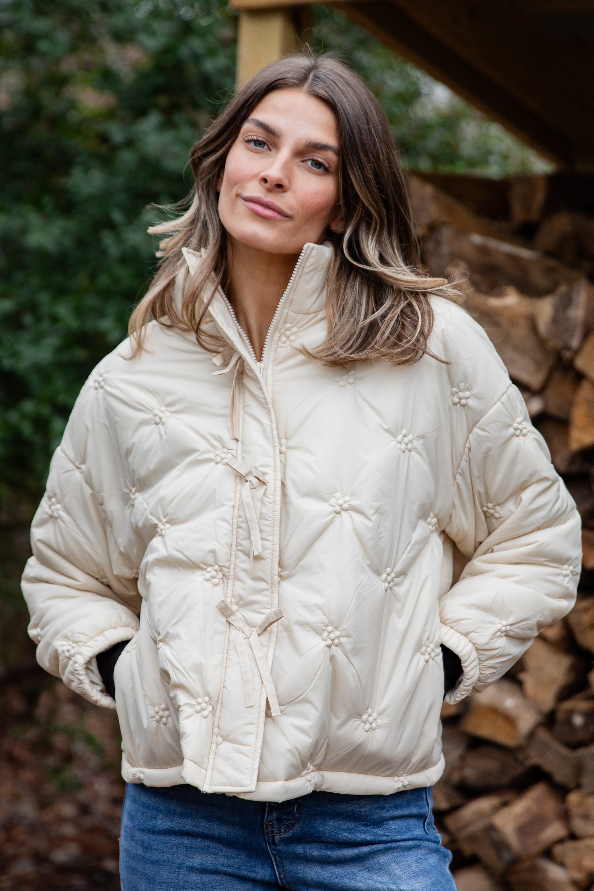Woman wearing a cream puffer jacket and blue jeans standing in front of stacked firewood.