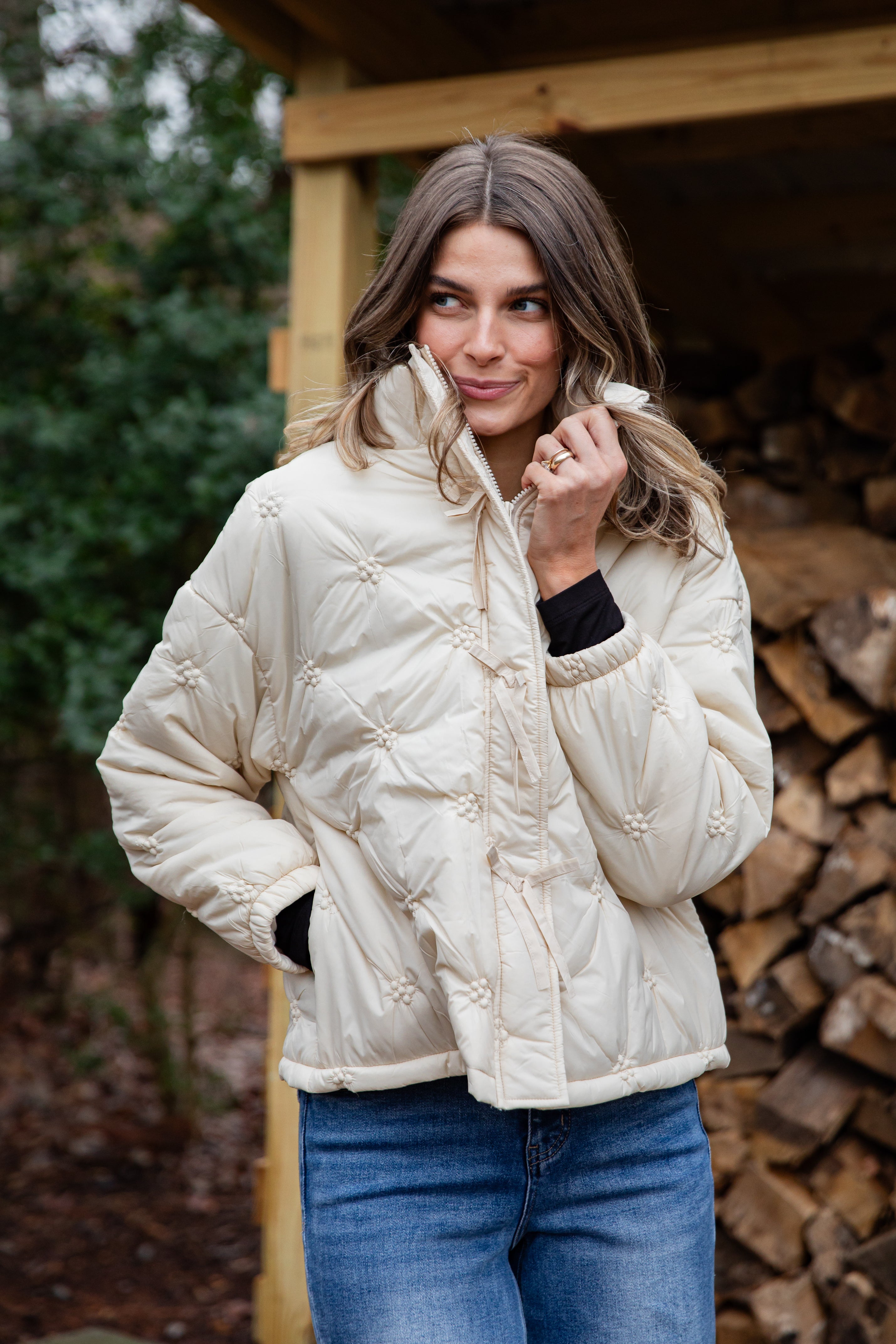 Woman wearing a cream quilted jacket standing in front of stacked firewood.