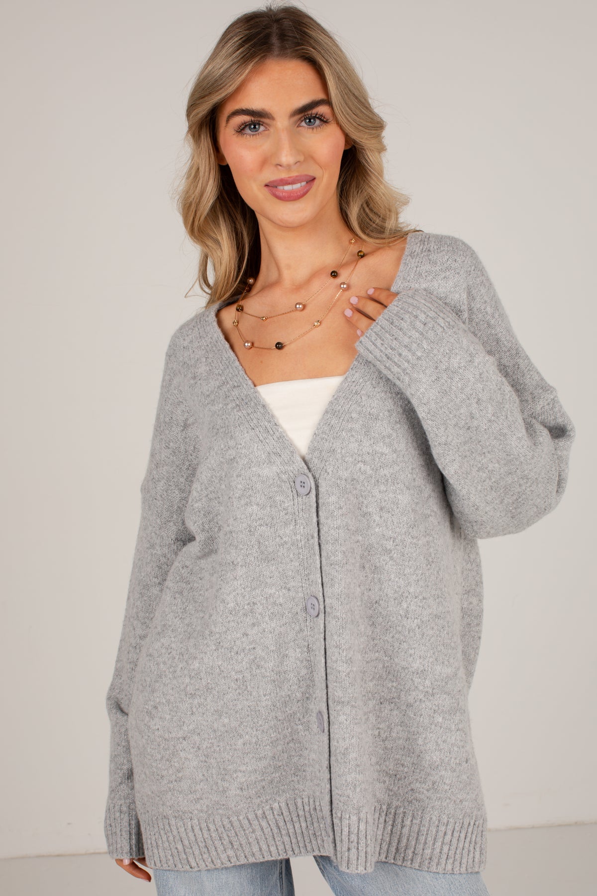 A woman wearing a light gray oversized knit cardigan with a V-neck and front buttons, layered over a white top and light blue jeans, against a plain light gray studio background.