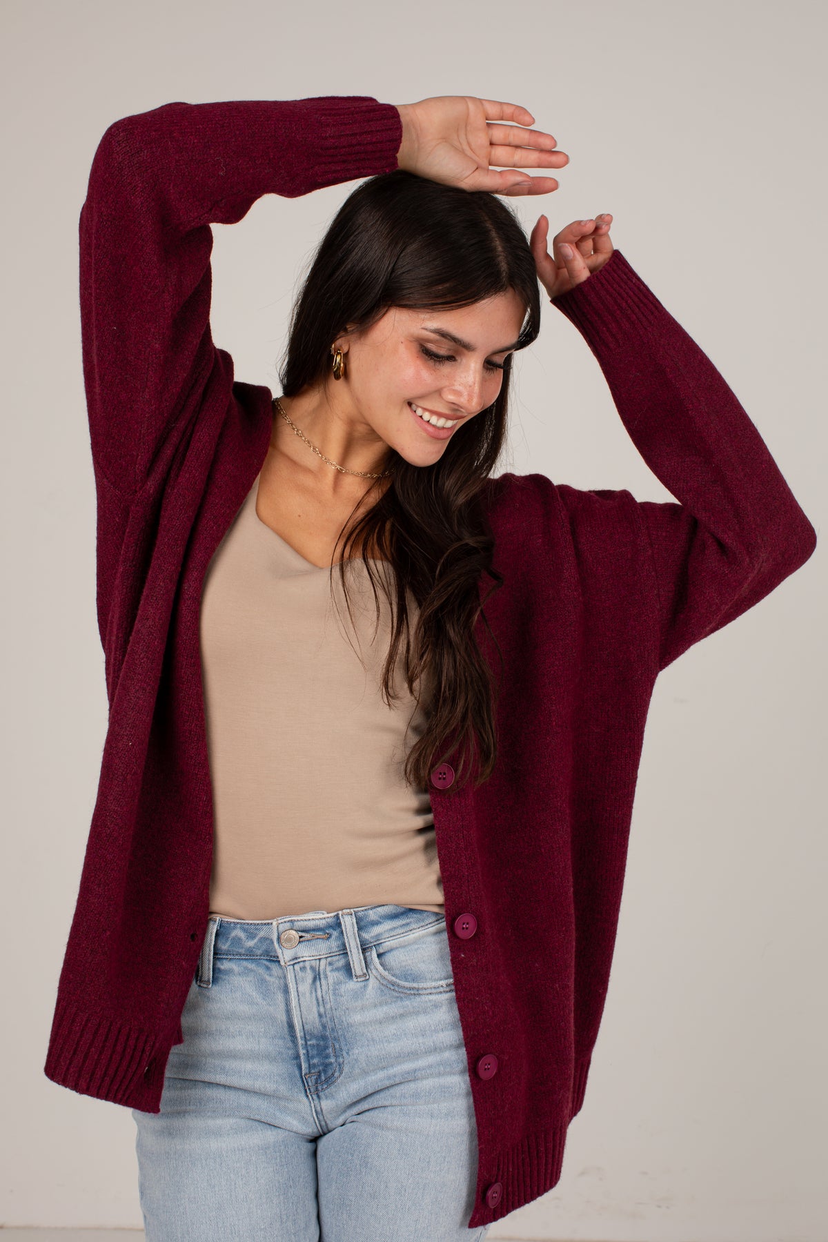 A woman wearing a burgundy knit cardigan over a beige top and light blue jeans, posing with her arms raised against a plain light gray background.