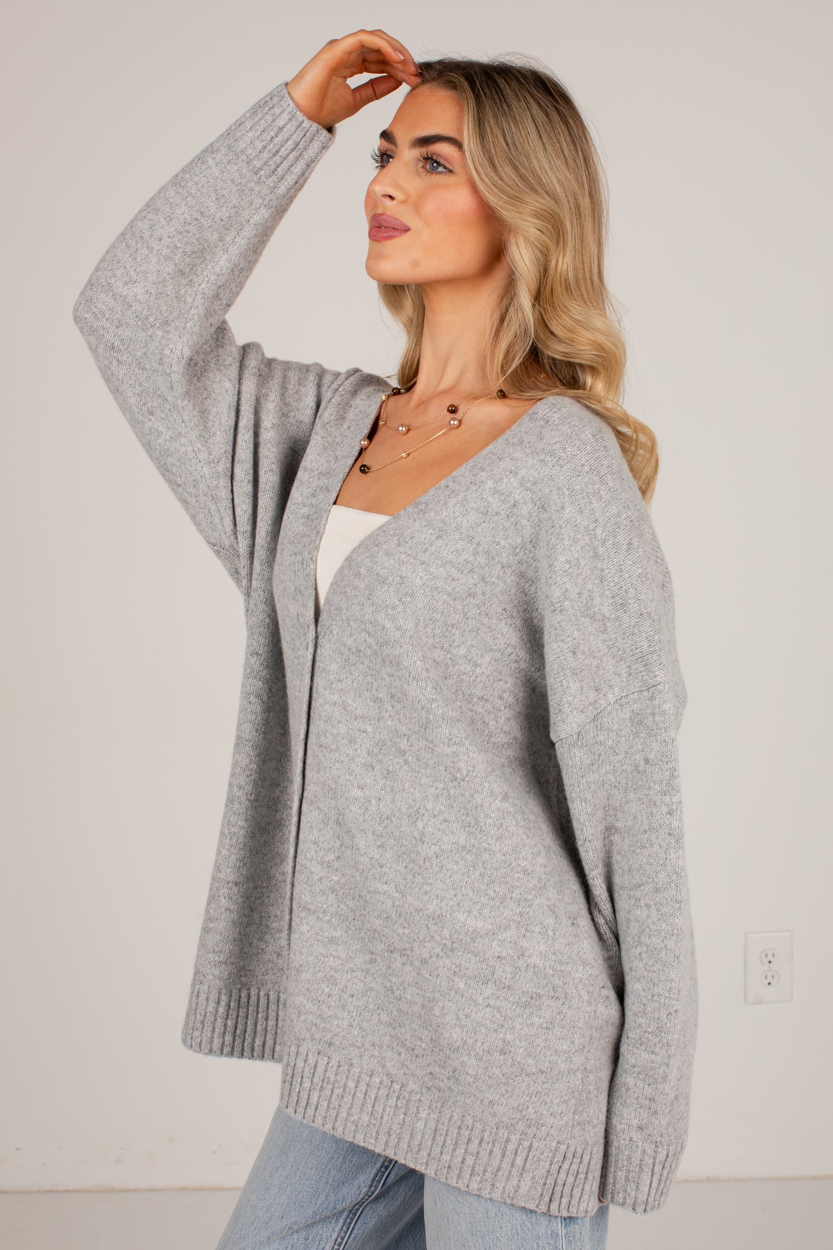 A woman wearing an oversized light gray knit cardigan over a white top and light blue jeans, standing against a plain off-white wall.