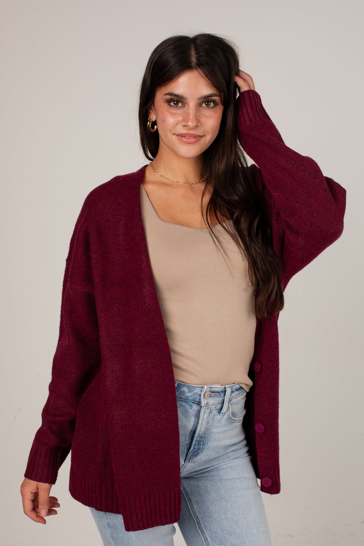 A woman modeling an open-front maroon knit cardigan over a beige tank top and light blue jeans, photographed against a plain light gray studio background.