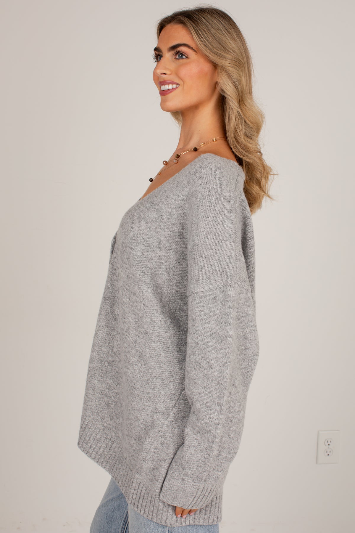 A woman wearing an oversized light gray knit sweater, shown in a side profile against a plain off-white wall.