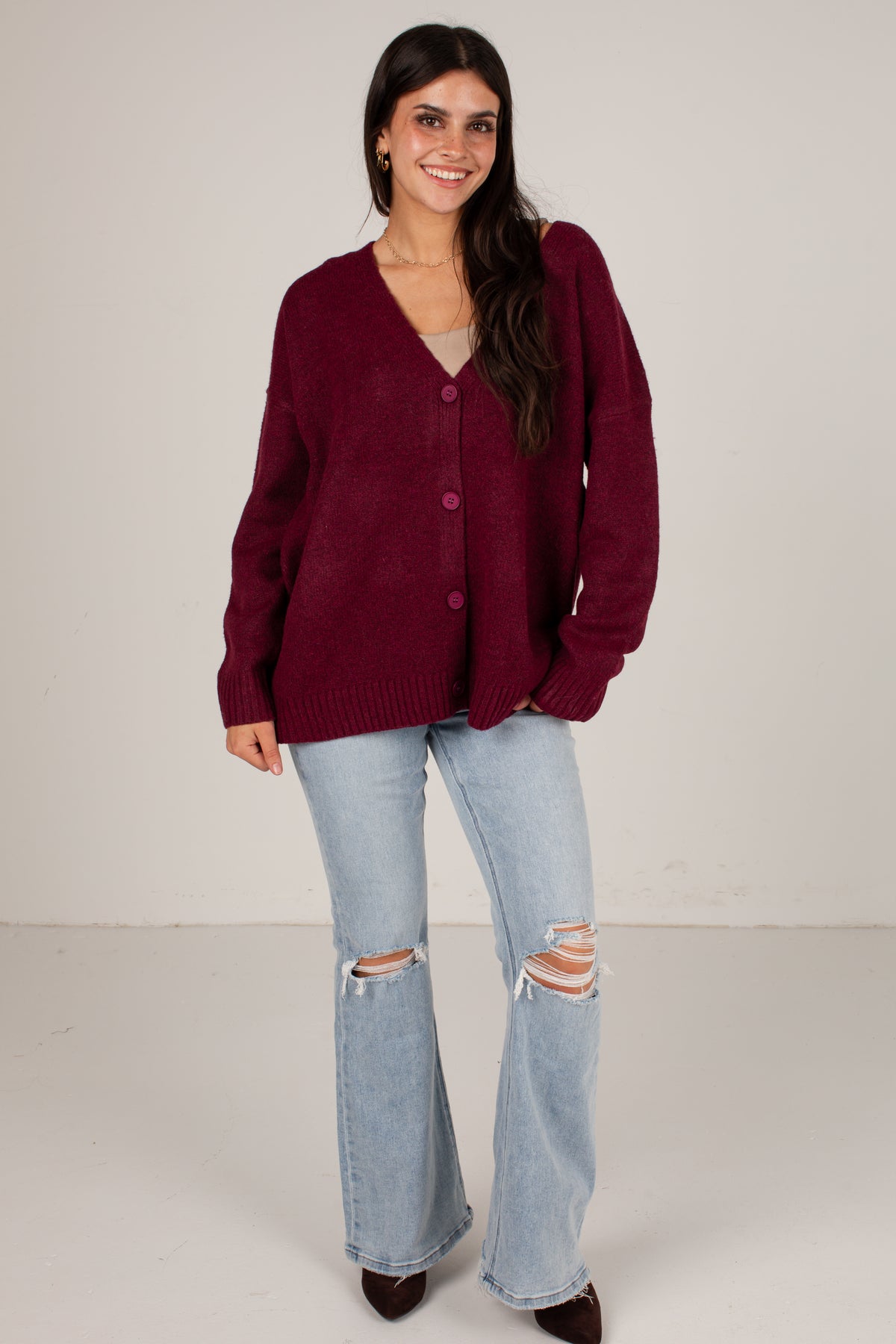 A woman modeling a maroon knit cardigan with a V-neck and button-front closure, paired with light-wash distressed flare jeans and dark brown pointed shoes, against a plain light gray studio background.