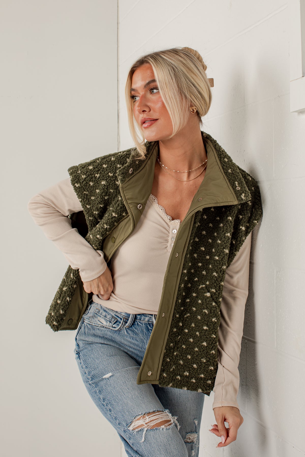 A woman wearing an olive green zip-front vest with a white floral pattern over a beige long-sleeve top and blue jeans, posing against a white wall.