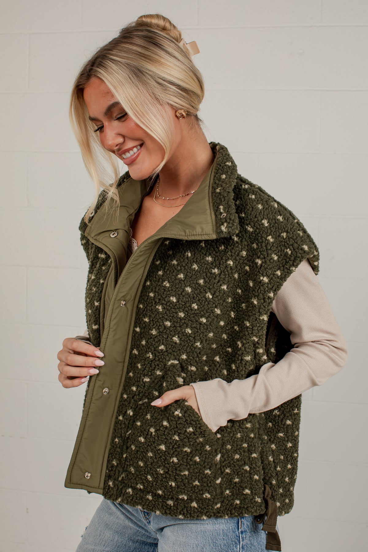 A woman wearing an olive green fleece vest with a beige and brown dalmatian-style spot pattern, layered over a light beige long-sleeve shirt and blue jeans, posing against a white brick wall.