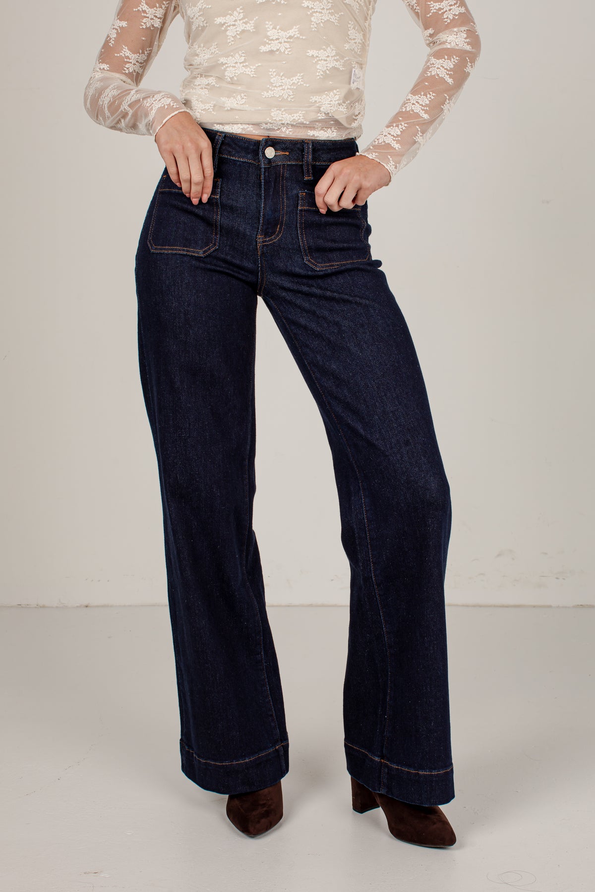 A model wearing dark blue wide-leg denim jeans with a cream lace long-sleeve top and brown pointed-toe heels, standing against a plain off-white studio background.