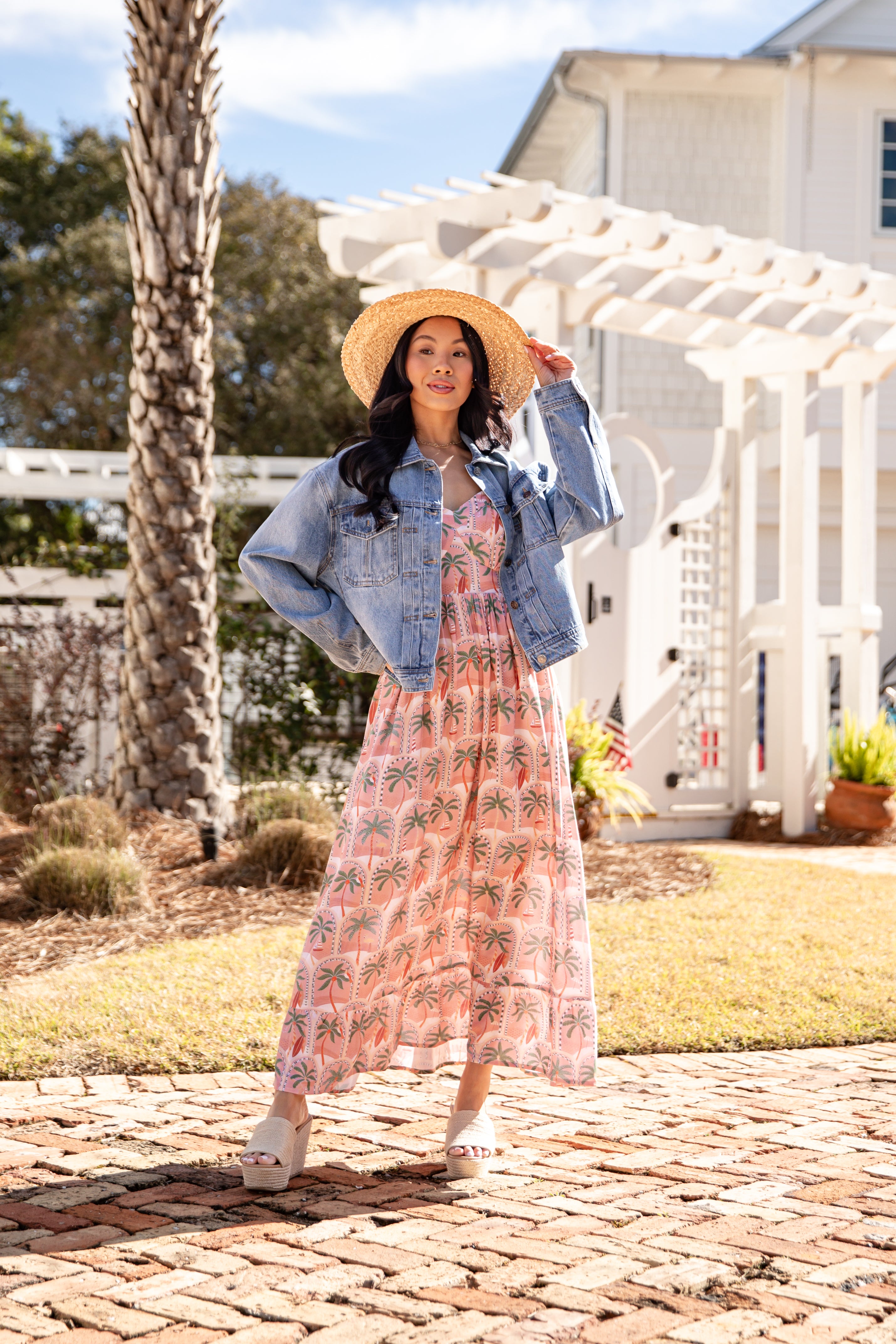 Woman in a floral dress and denim jacket standing on a brick path with a white building and palm tree in the background.