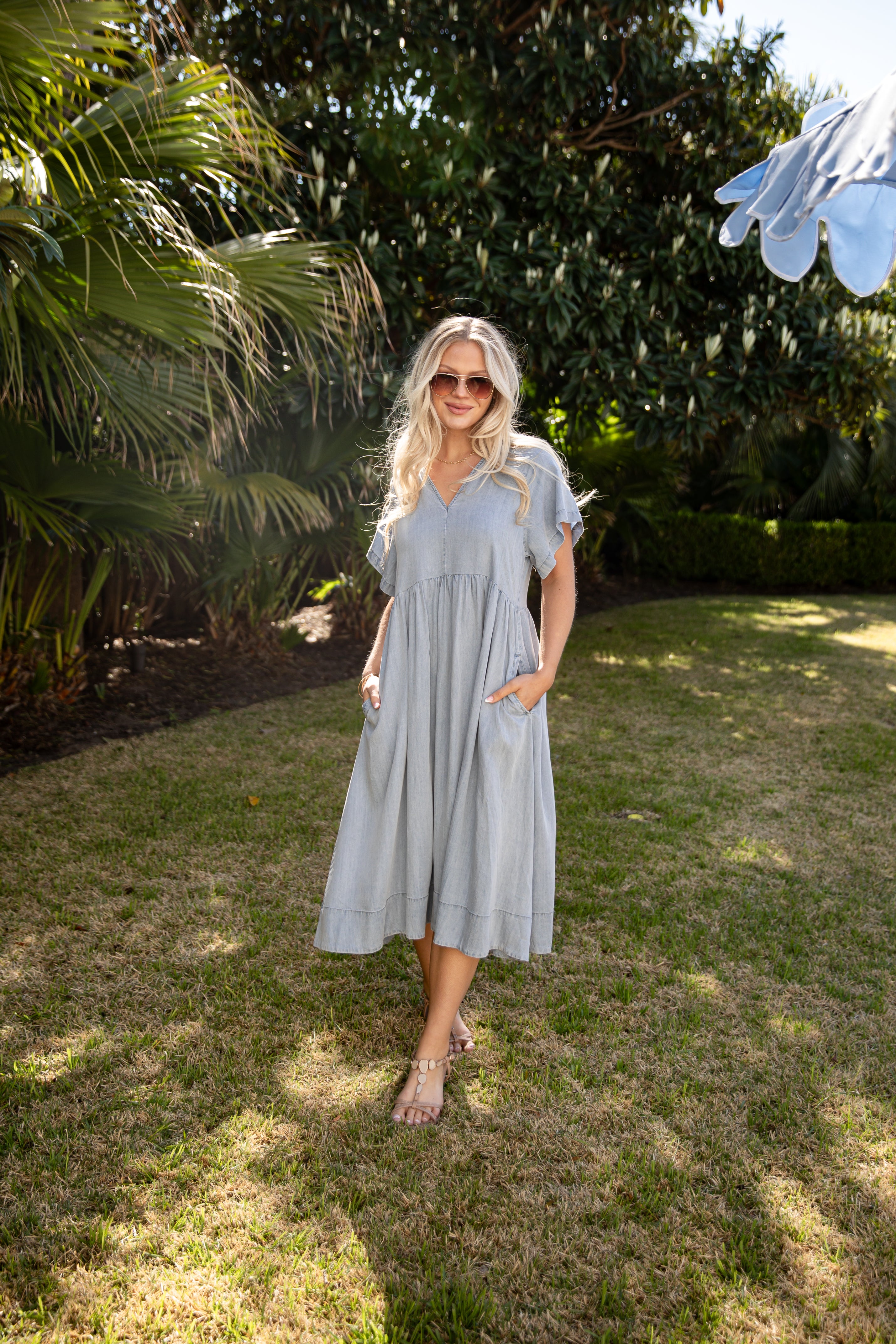 Woman in a light blue dress standing in a garden with trees and grass.