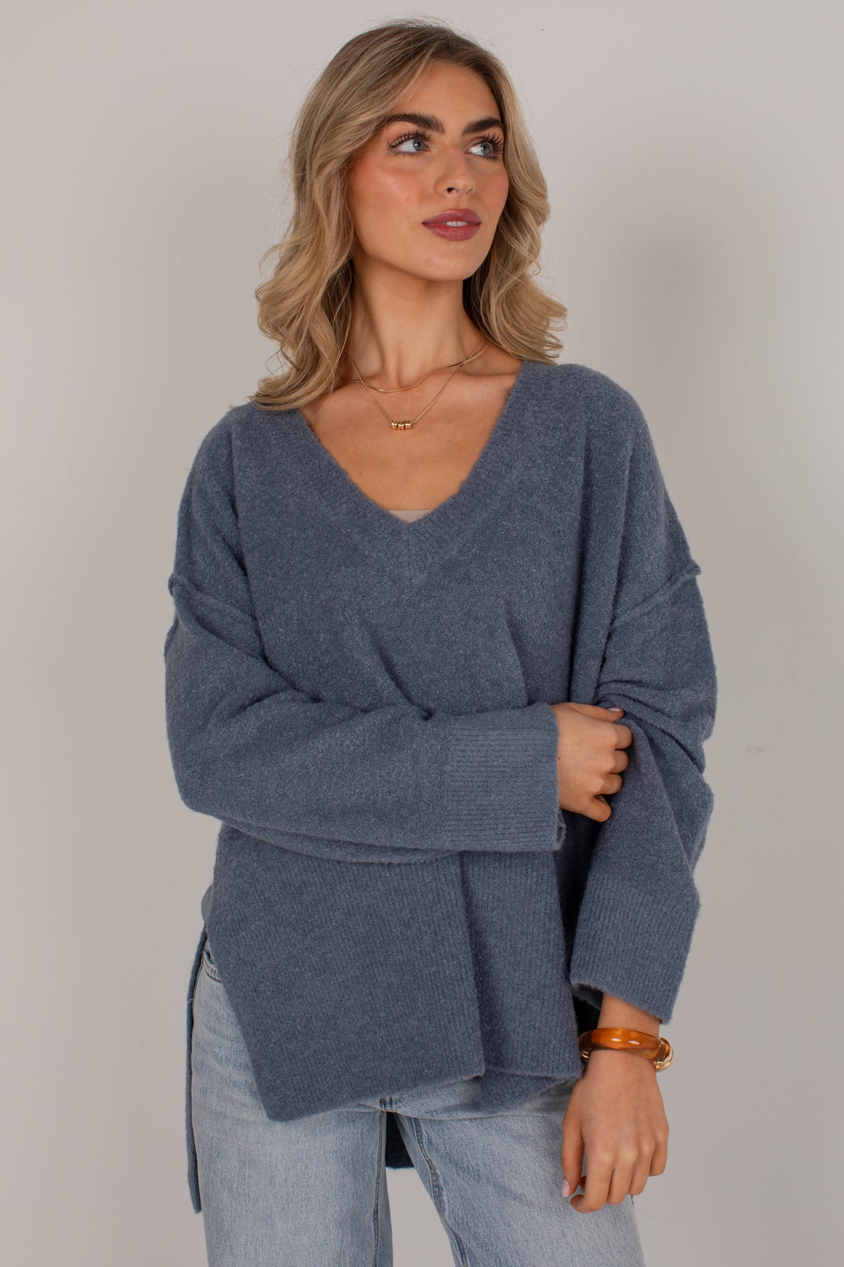 A woman wearing a slate blue knit sweater with a V-neck and wide sleeves, paired with light-wash jeans, photographed against a plain light gray background.