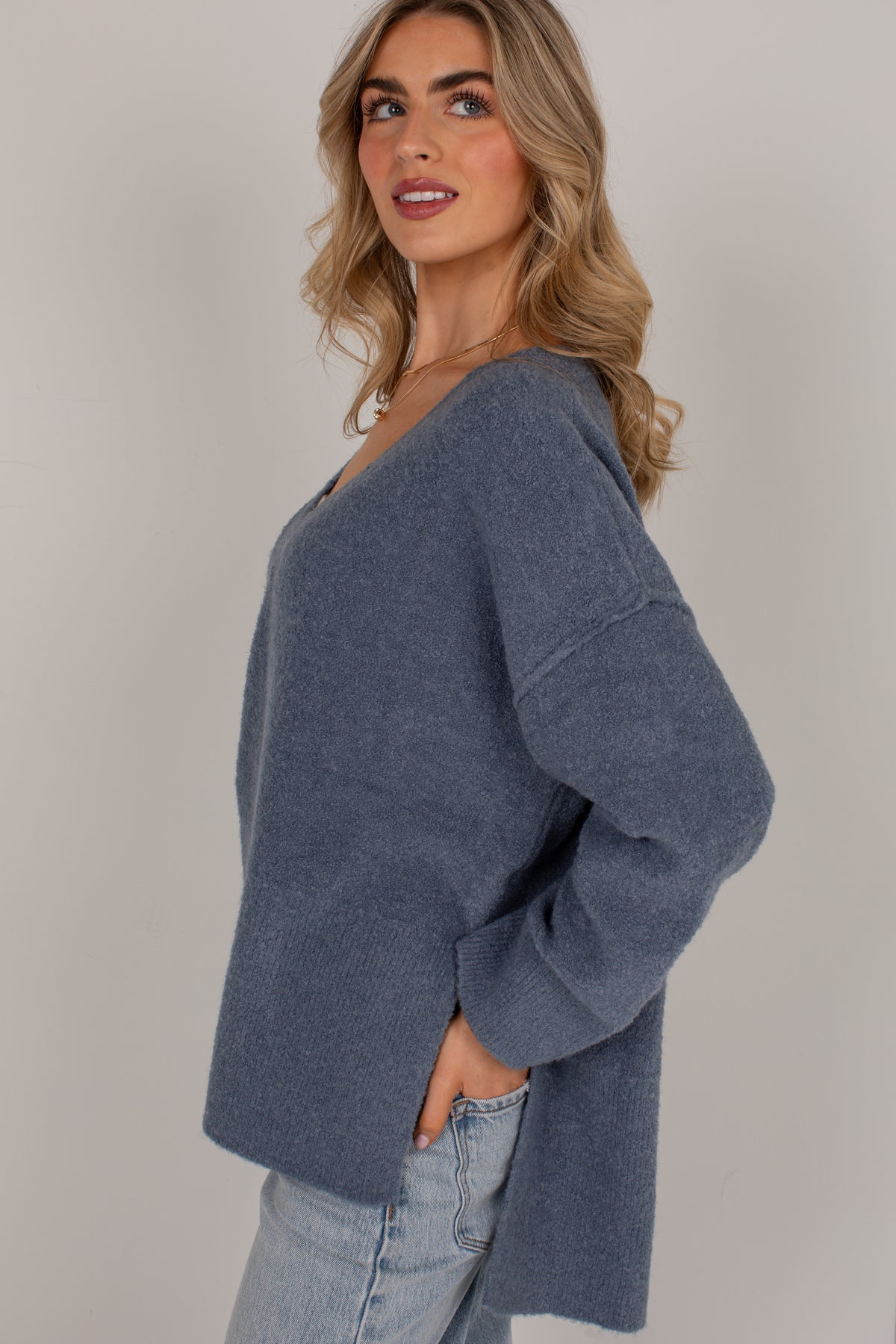 A woman wearing a loose, long-sleeve knit sweater in a muted slate blue-gray color, paired with light-wash jeans, photographed against a plain light gray studio background.