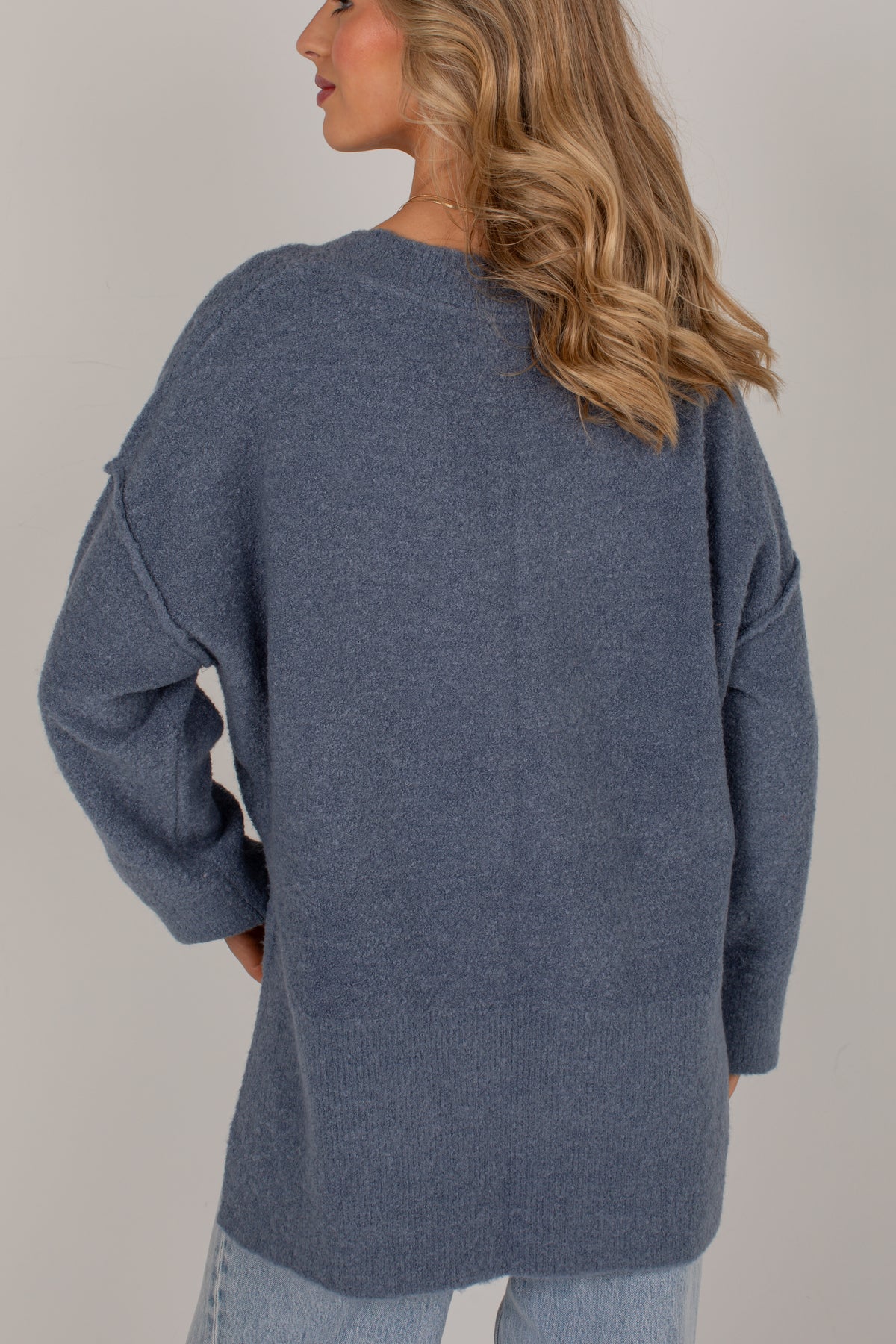 A woman wearing an oversized heathered slate blue knit sweater, shown from the back against a plain light gray studio background.
