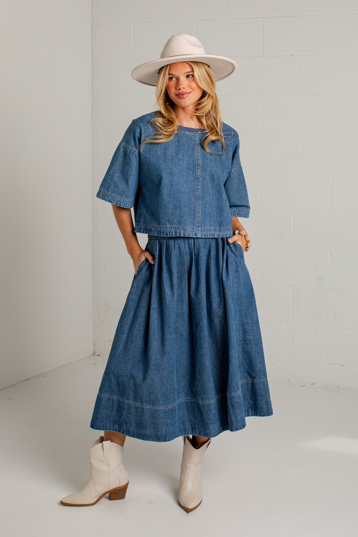 A woman modeling a blue denim two-piece set consisting of a short-sleeve cropped top and a matching midi skirt, styled with a cream wide-brim hat and cream ankle boots against a white brick wall background.