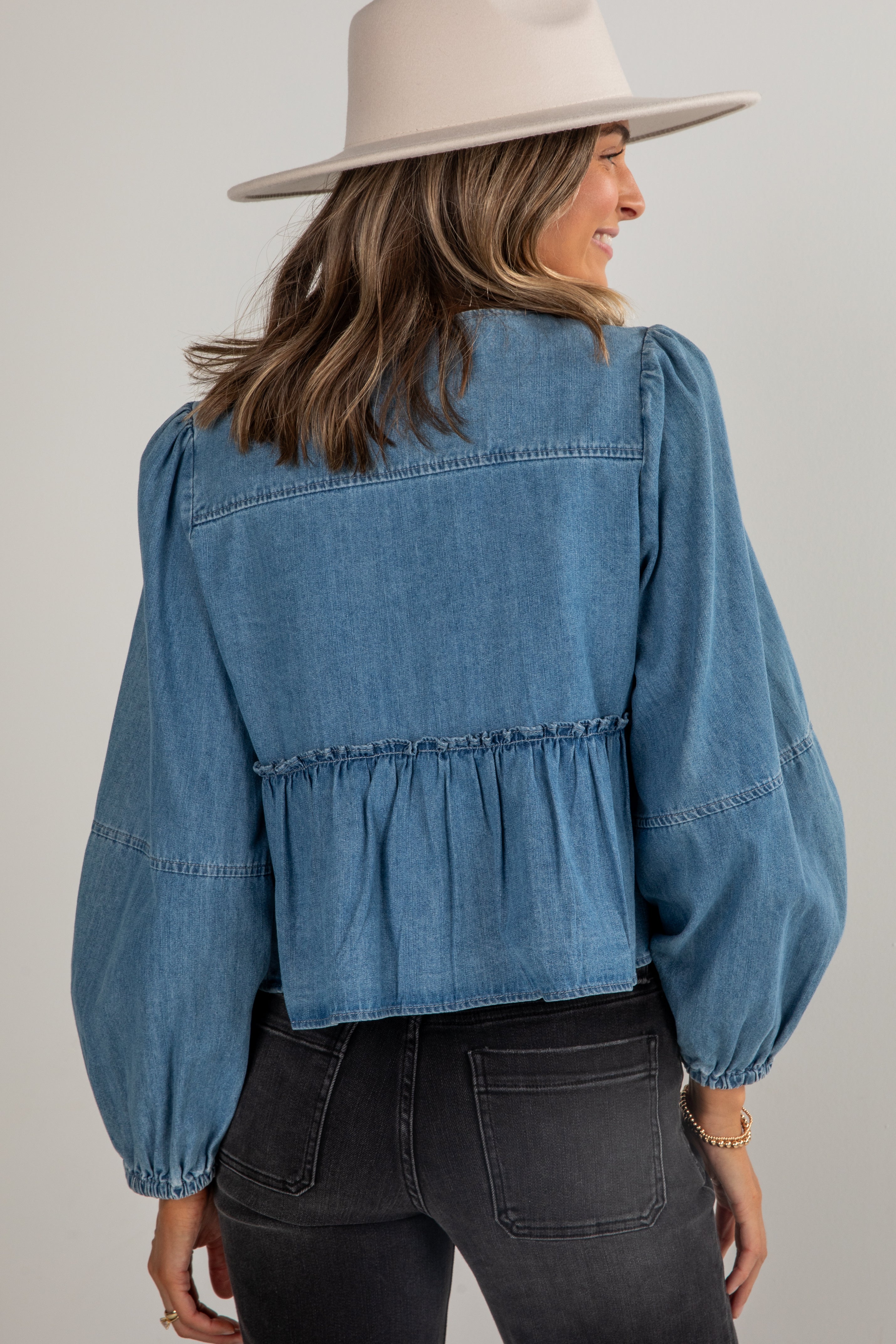 Woman wearing a blue denim blouse with ruffled details and a beige hat on a plain background