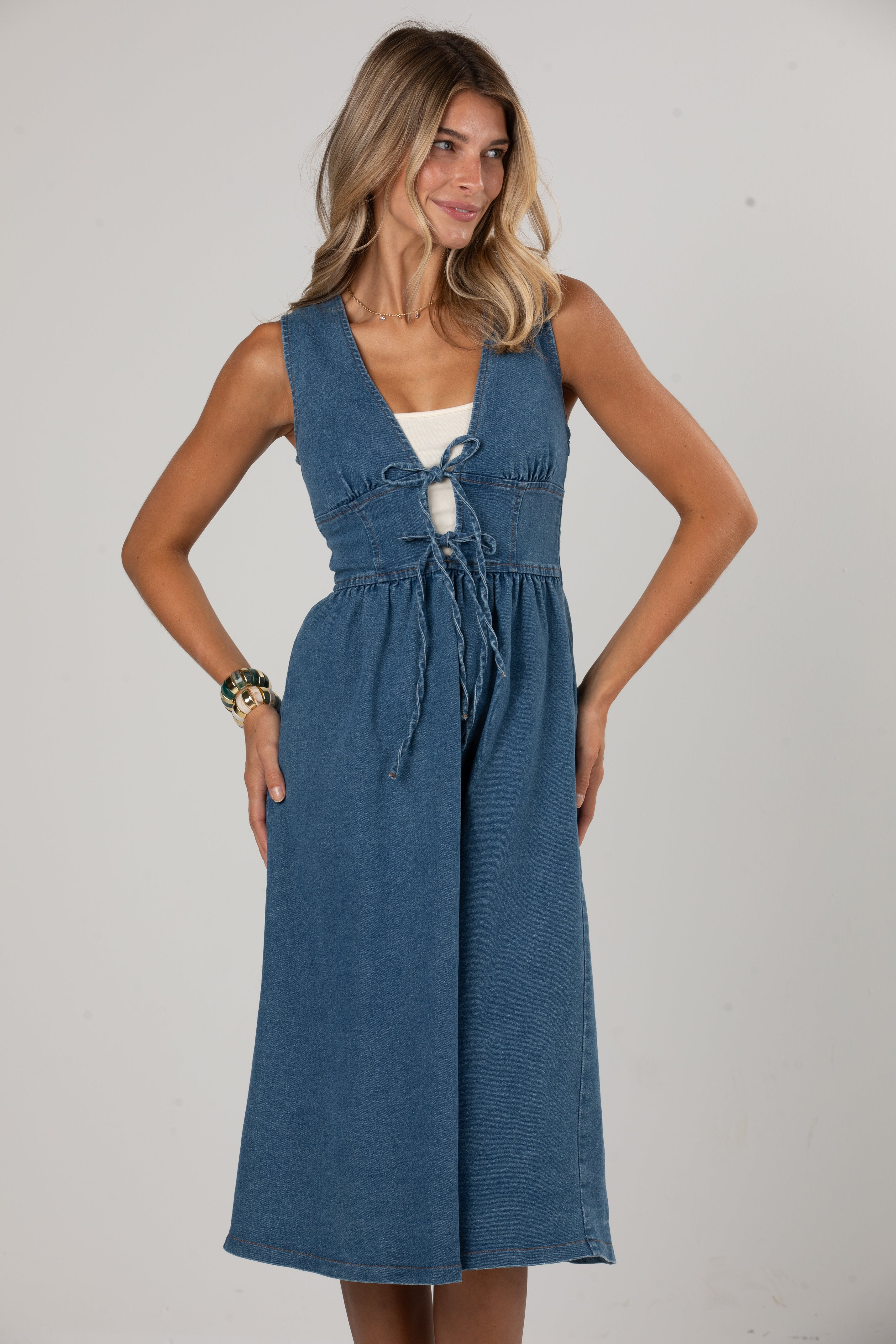 A woman modeling a sleeveless blue denim midi dress with a tie-front detail over a white underlayer, photographed against a plain light gray studio background.