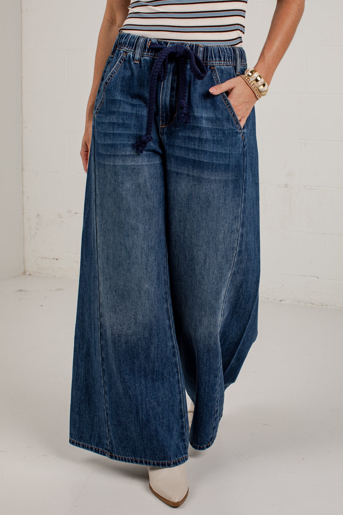 A person wearing wide-leg blue denim jeans with a dark drawstring waist, paired with a striped top and white pointed shoes, standing against a white studio wall.