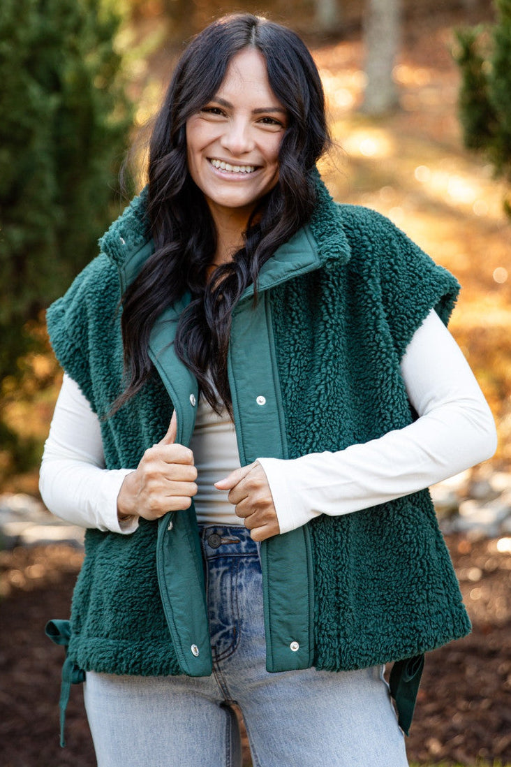A woman wearing a dark green sherpa fleece vest over a white long-sleeve shirt and light blue jeans, standing outdoors in a park-like setting with trees and autumn foliage in the background.