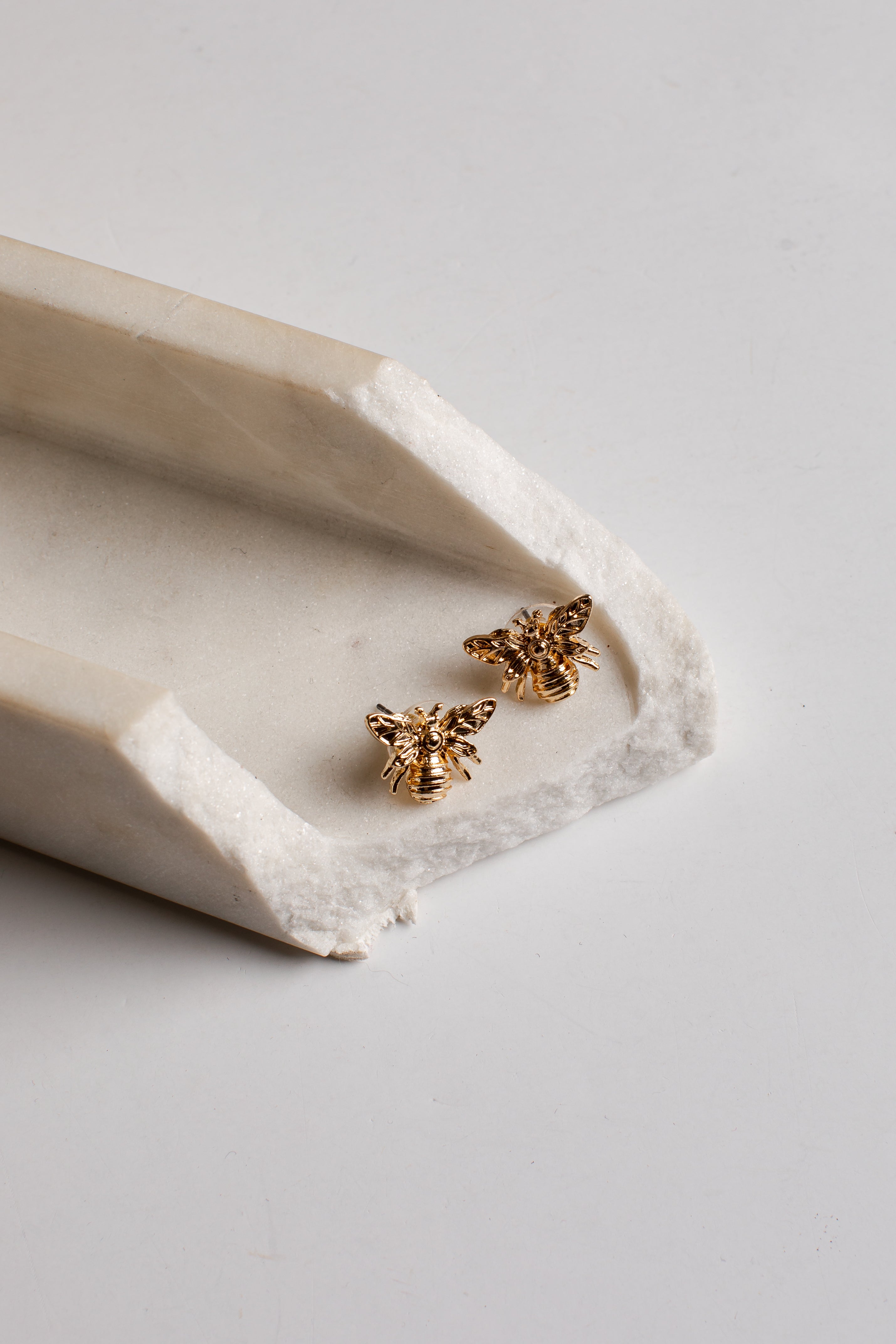 A pair of small gold bee stud earrings resting on a white ceramic tray against a light gray background.