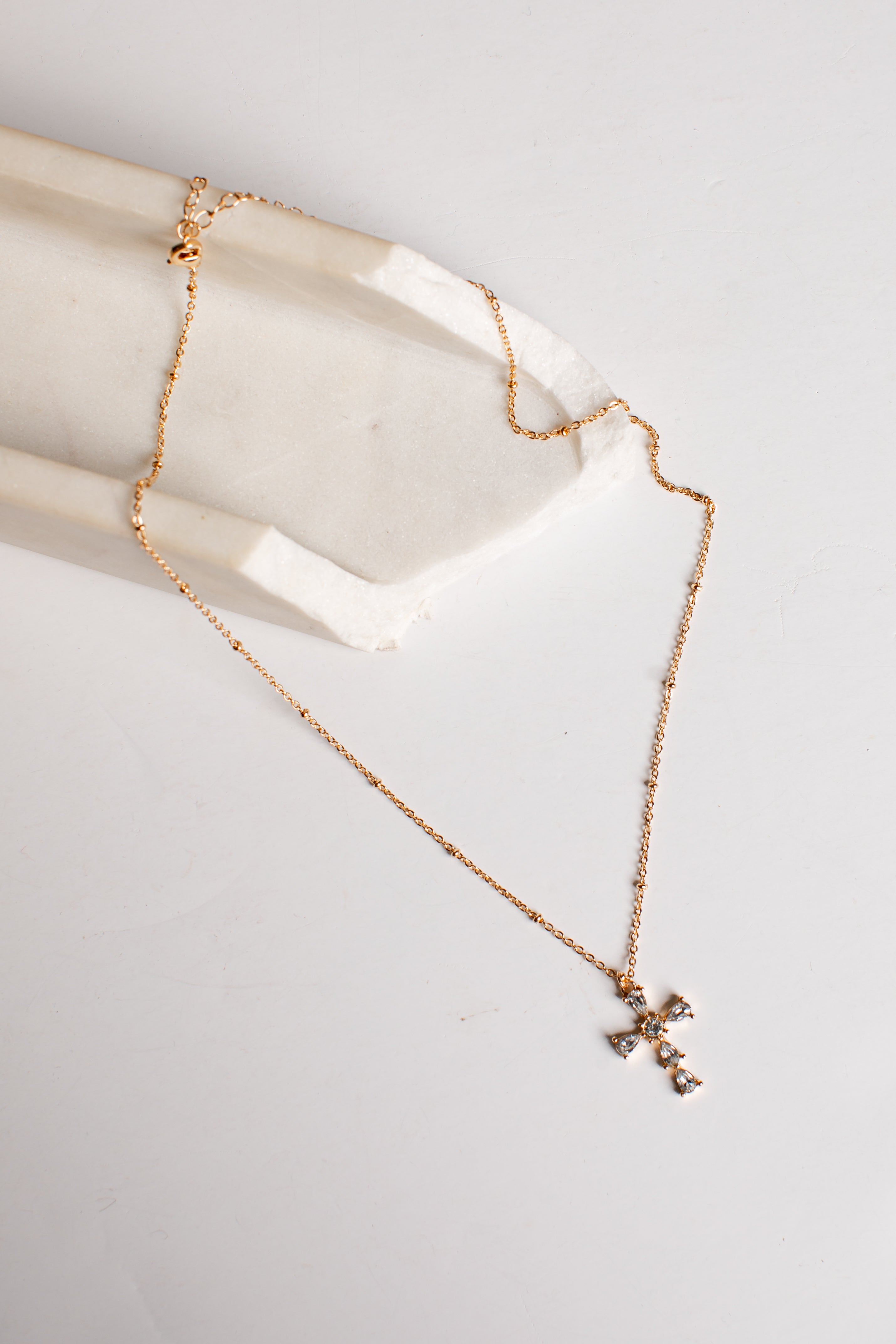 A delicate rose gold chain necklace with a small cross pendant, draped over a white stone display block on a light gray background.