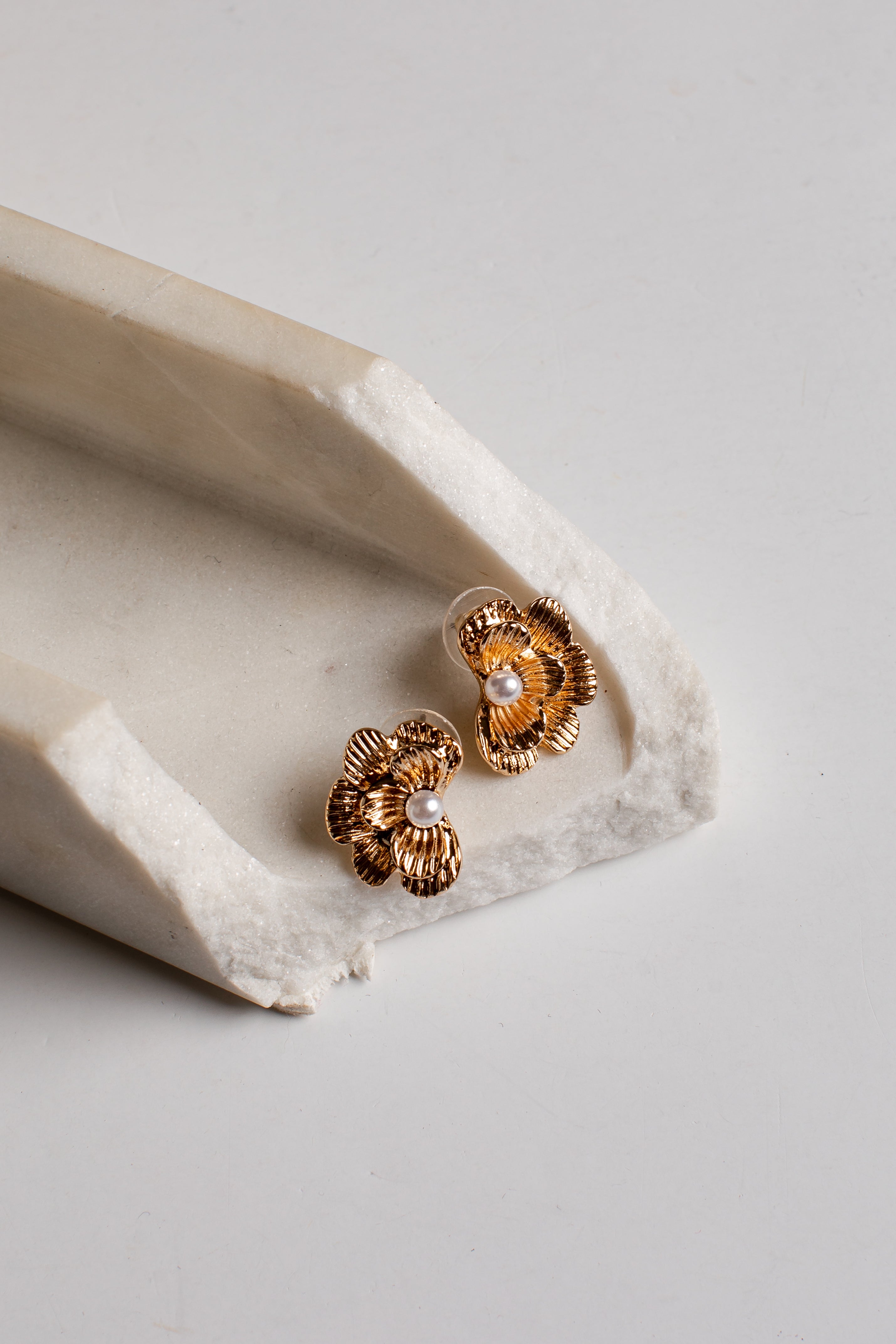 A pair of gold-toned flower-shaped stud earrings with small white pearl centers, resting on a textured off-white stone tray against a light gray background.