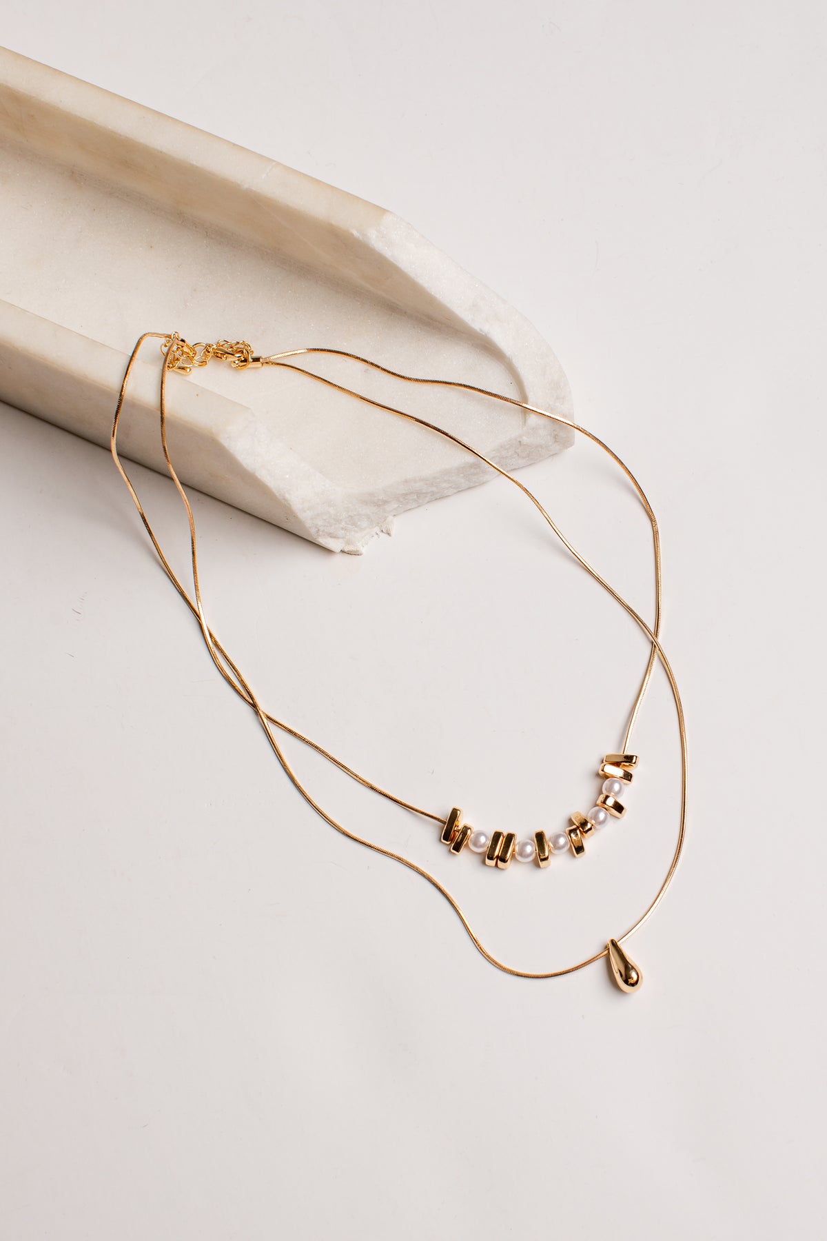 A delicate gold double-layer necklace with a small cluster of clear rhinestones and a tiny teardrop charm, displayed on a light beige textured ceramic prop against a white background.