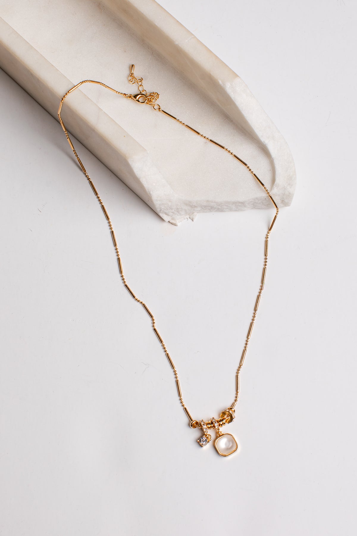 A delicate gold chain necklace with a small charm featuring a clear gemstone and a gold square pendant, displayed on a light beige stone tray against a white background.
