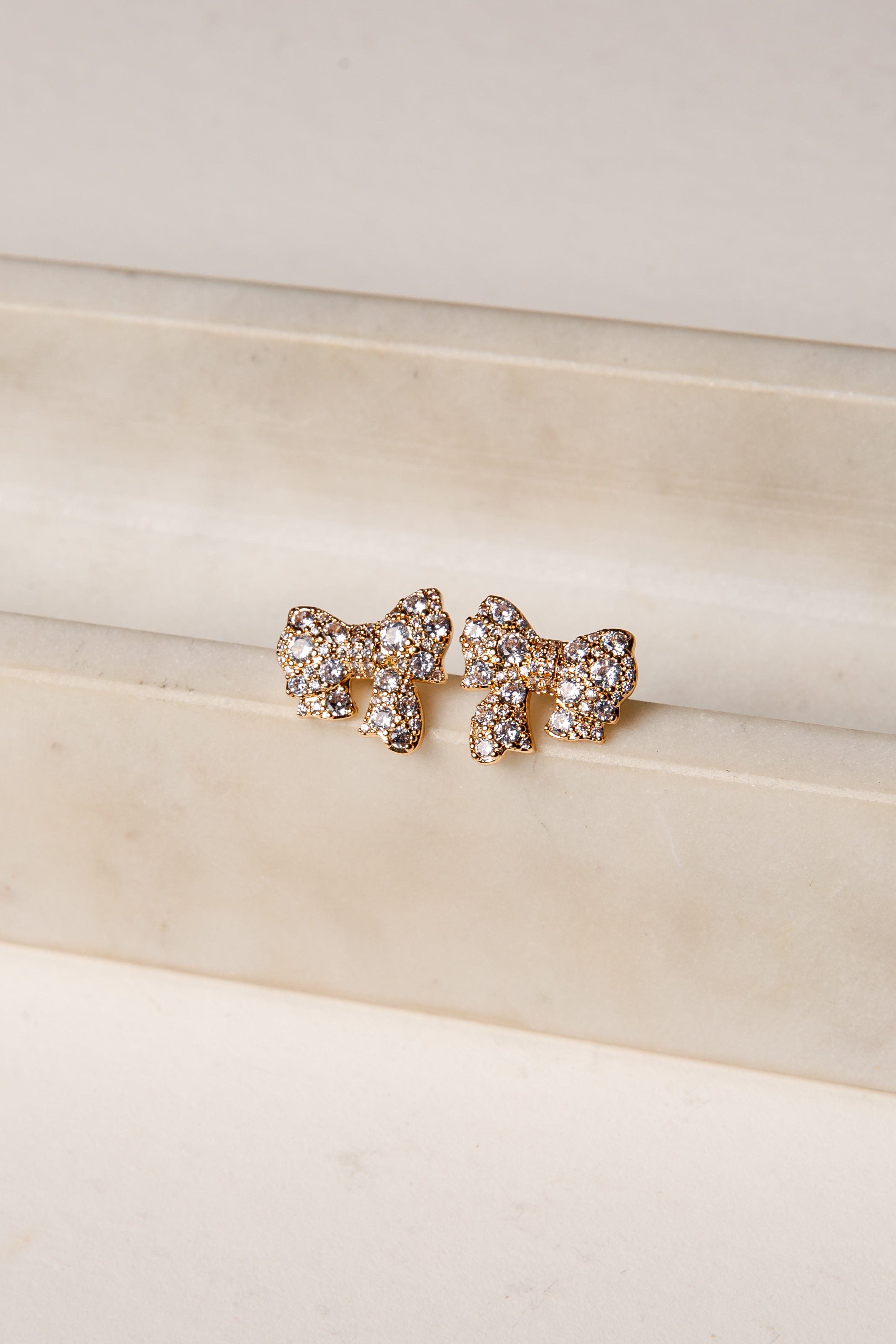 Gold bow-shaped earrings with rhinestones on a beige surface