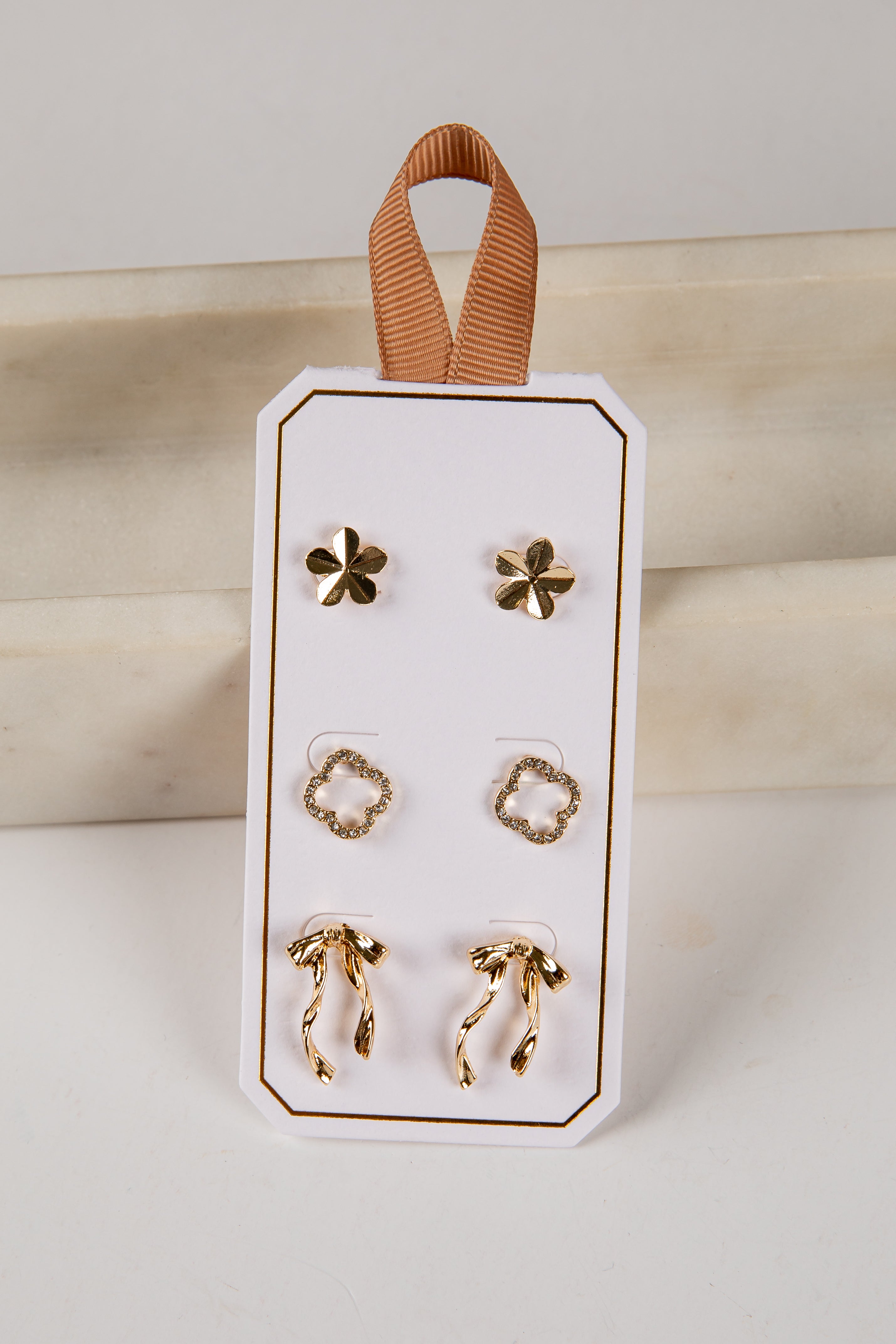 Set of gold earrings on a white card with a brown ribbon, against a neutral background.