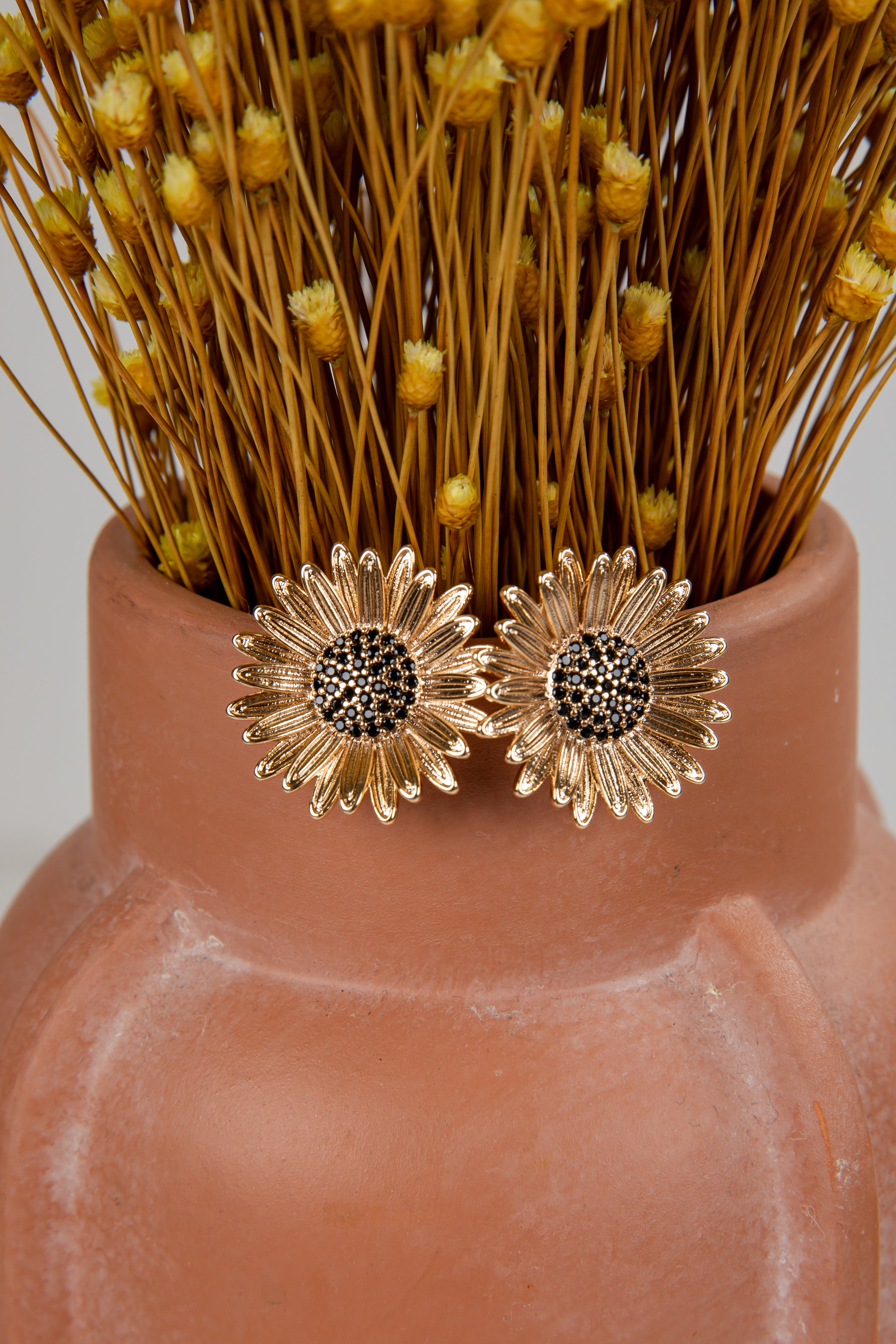 Gold sunflower earrings on a terracotta vase with dried flowers.