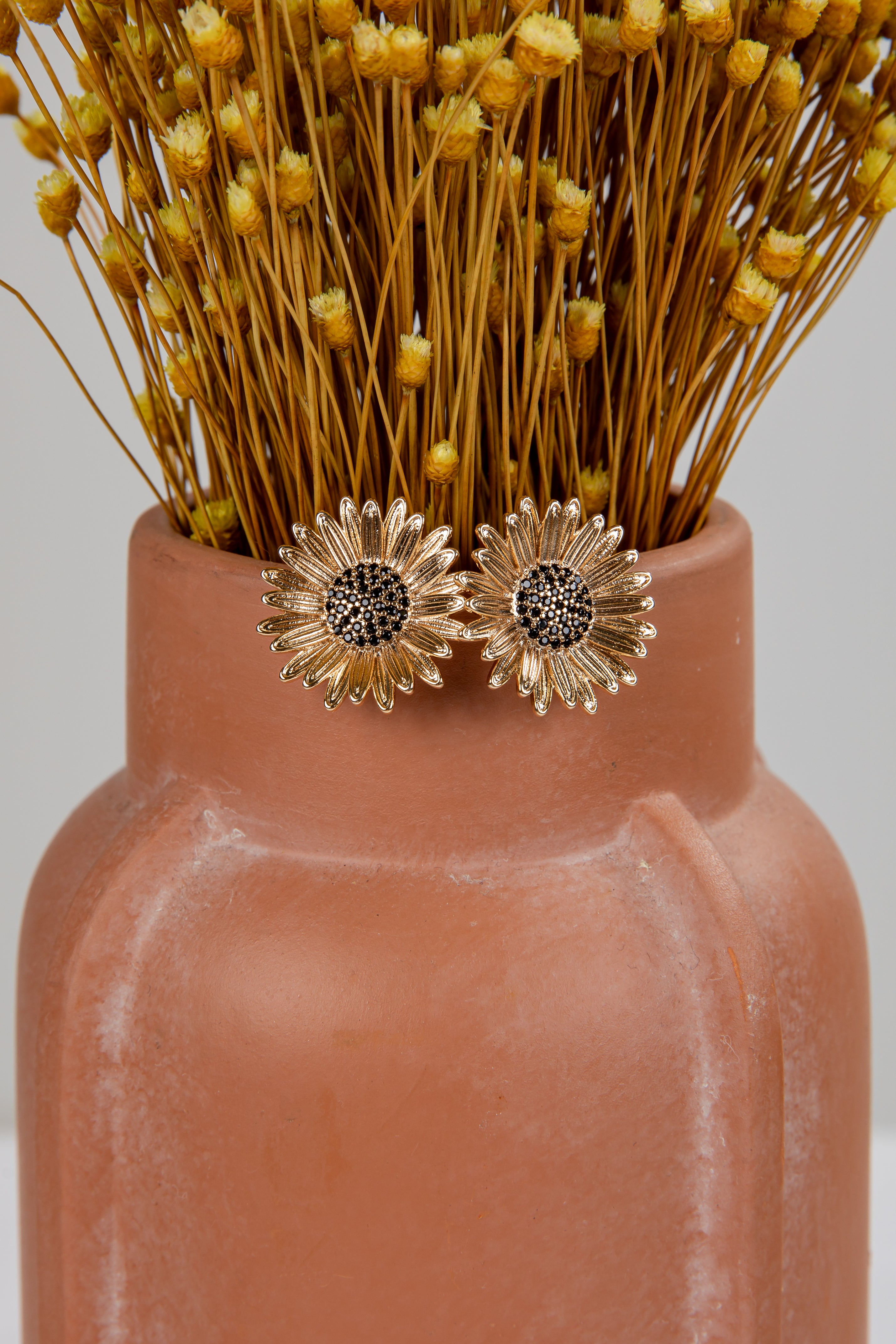 Gold sunflower earrings on a terracotta vase with dried flowers.