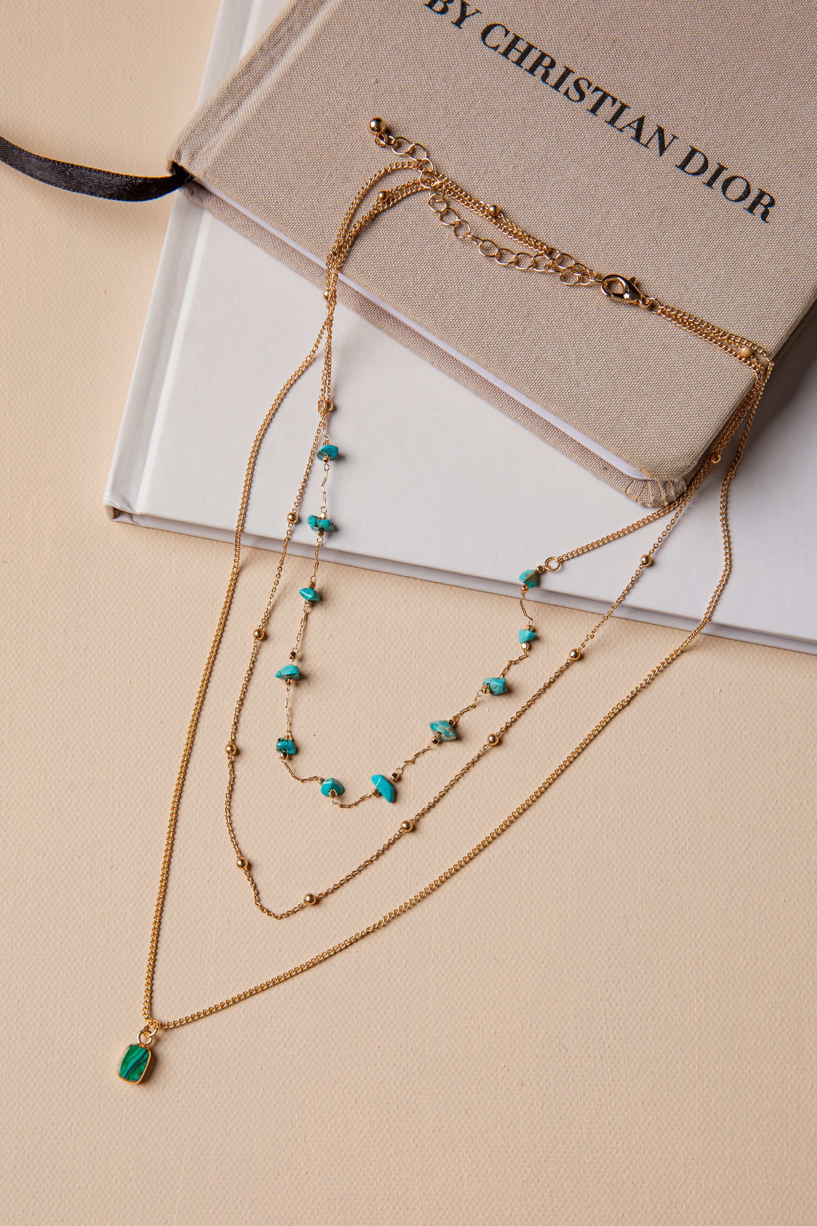 Layered gold necklace with green and blue stones on a beige surface with a Christian Dior book.