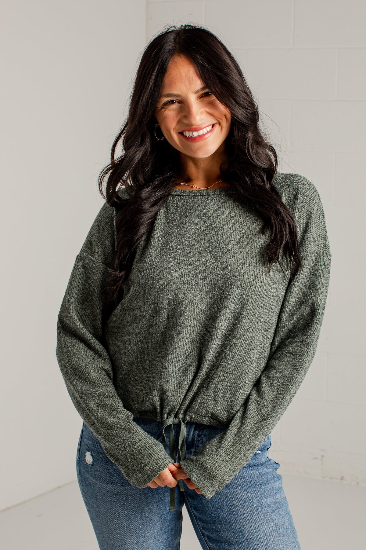 A woman modeling a loose, heathered sage green knit sweater with a tied hem, paired with blue jeans and brown heeled sandals, standing against a white studio wall.