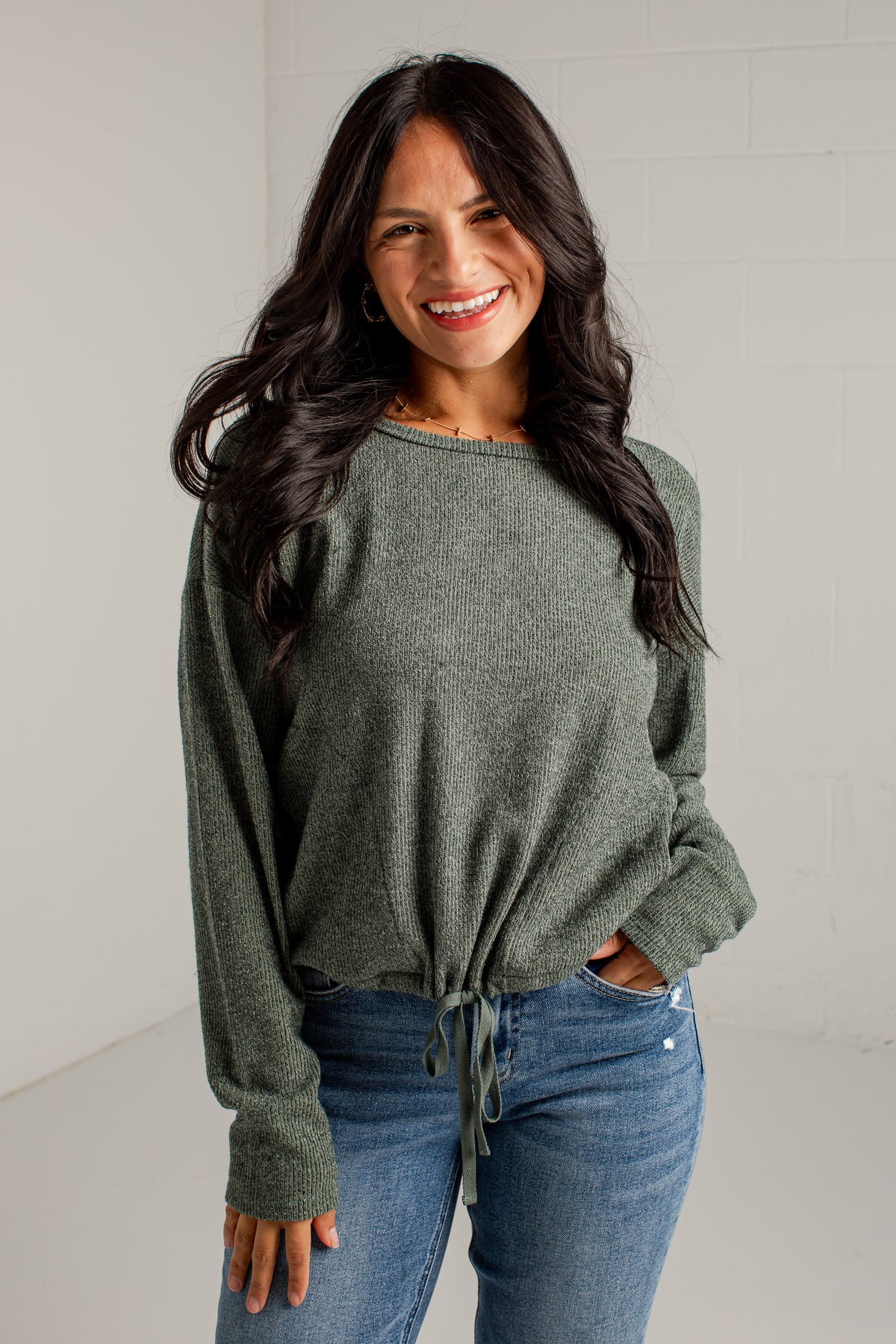 A woman modeling a loose, heathered sage green knit sweater with a tied hem, paired with blue jeans and brown heeled sandals, standing against a white studio wall.