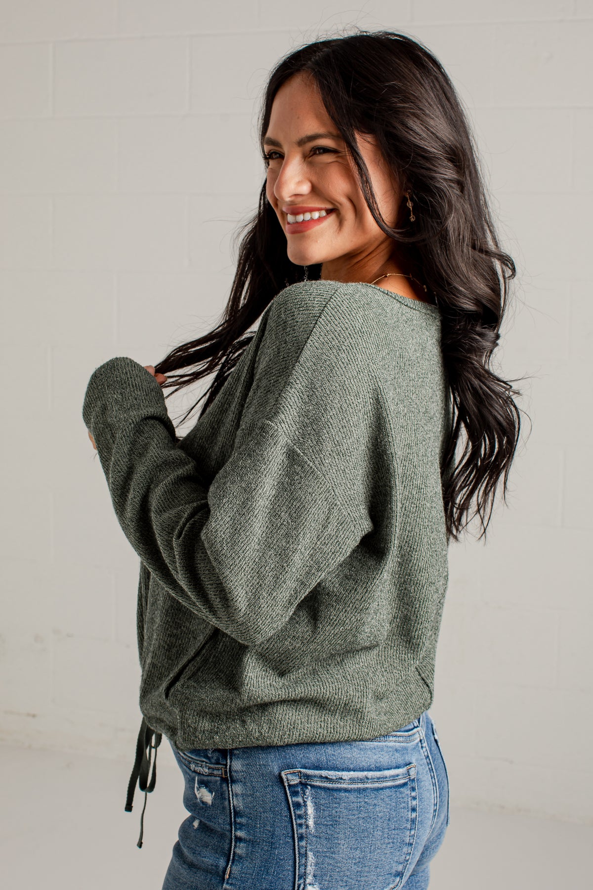 A woman with long dark hair is shown from the back and side, wearing a textured sage green knit sweater and blue jeans against a light gray brick wall.