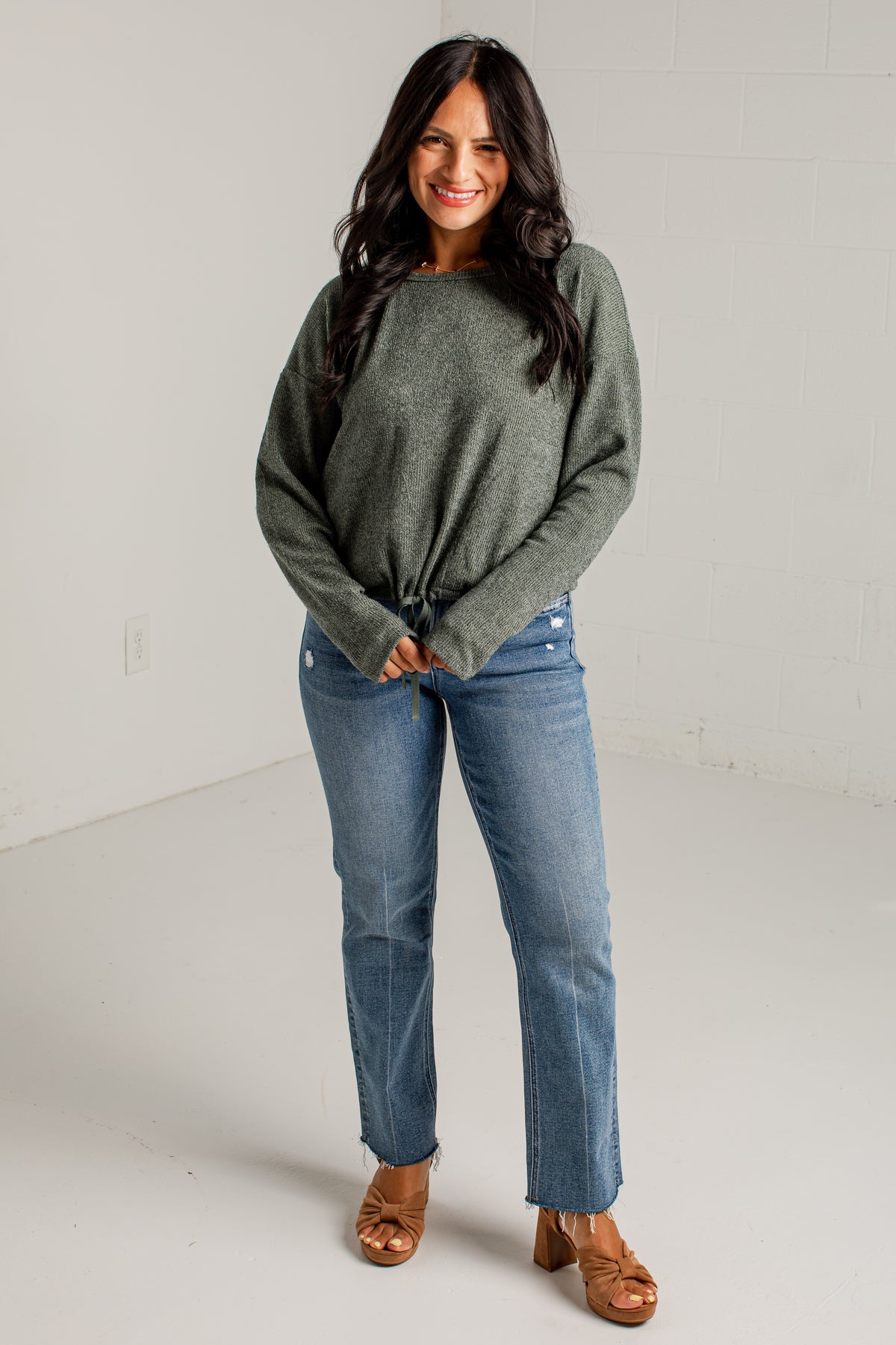 A woman modeling a loose, heathered sage green knit sweater with a tied hem, paired with blue jeans and brown heeled sandals, standing against a white studio wall.