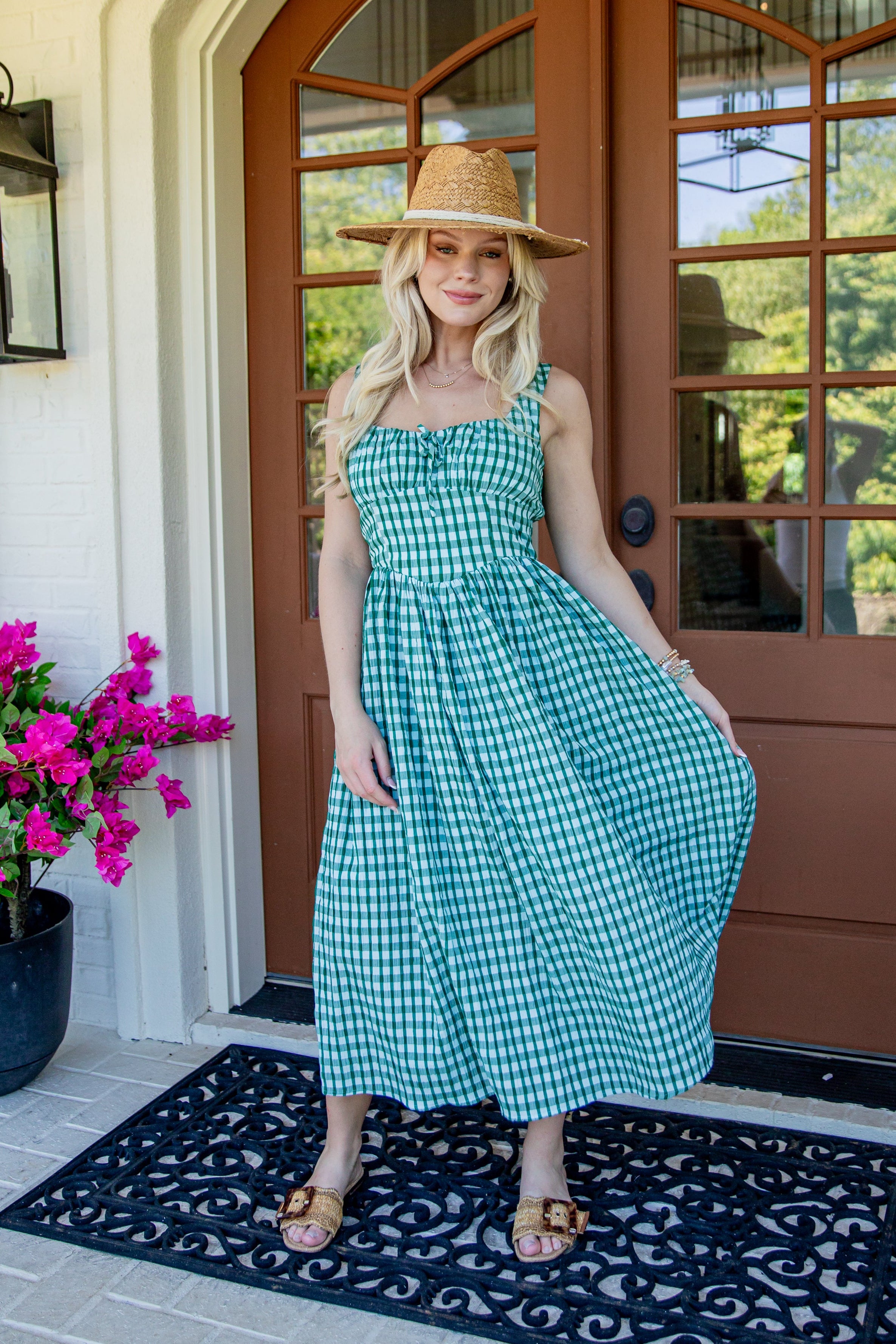 A woman wearing a green and white gingham midi dress, a straw hat, and sandals stands on a black patterned doormat in front of a brown wooden front door with a potted plant on the left.