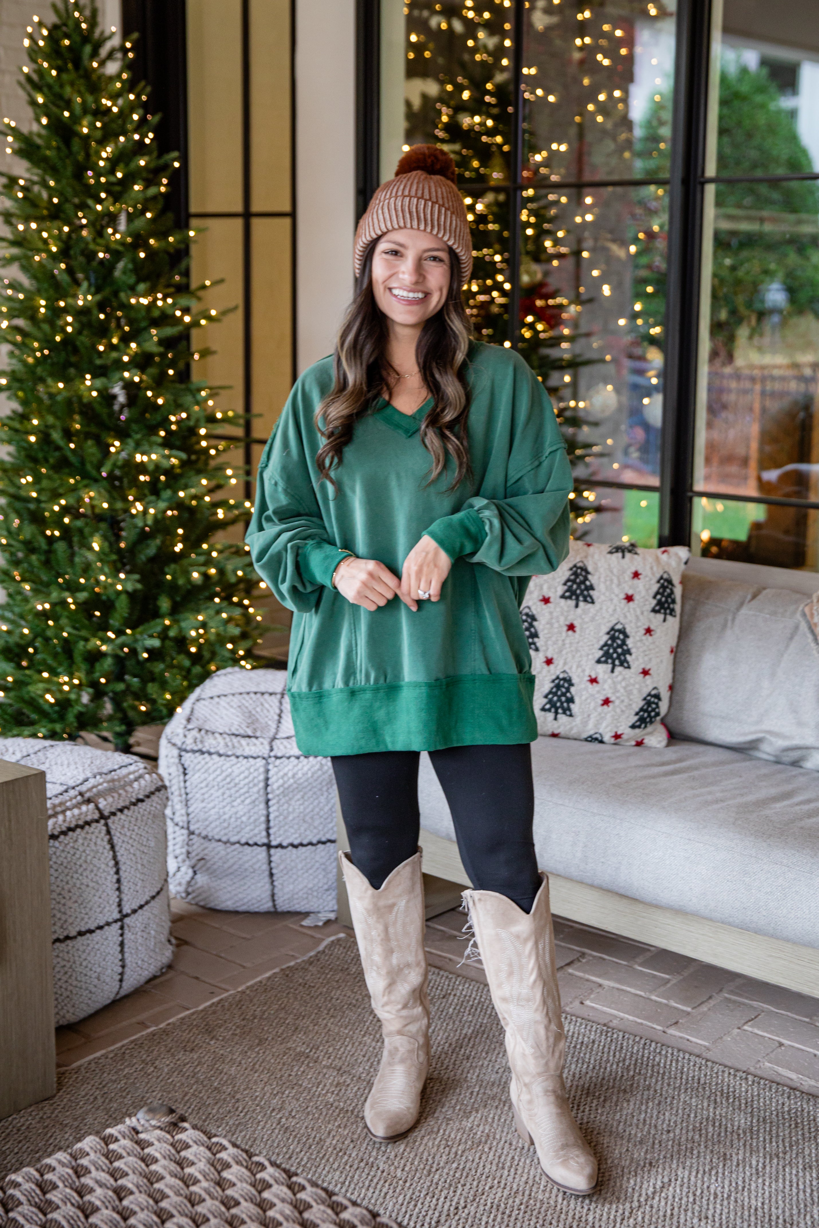 Woman in a green sweater and beige boots standing in a living room with Christmas decorations.