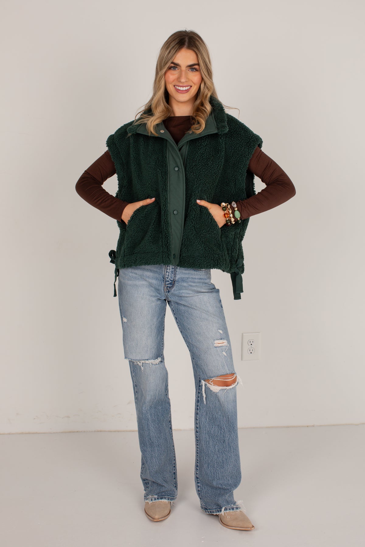 A woman modeling a dark forest green fleece vest over a brown long-sleeve top, paired with light-wash distressed jeans and tan boots, standing against a plain off-white wall.