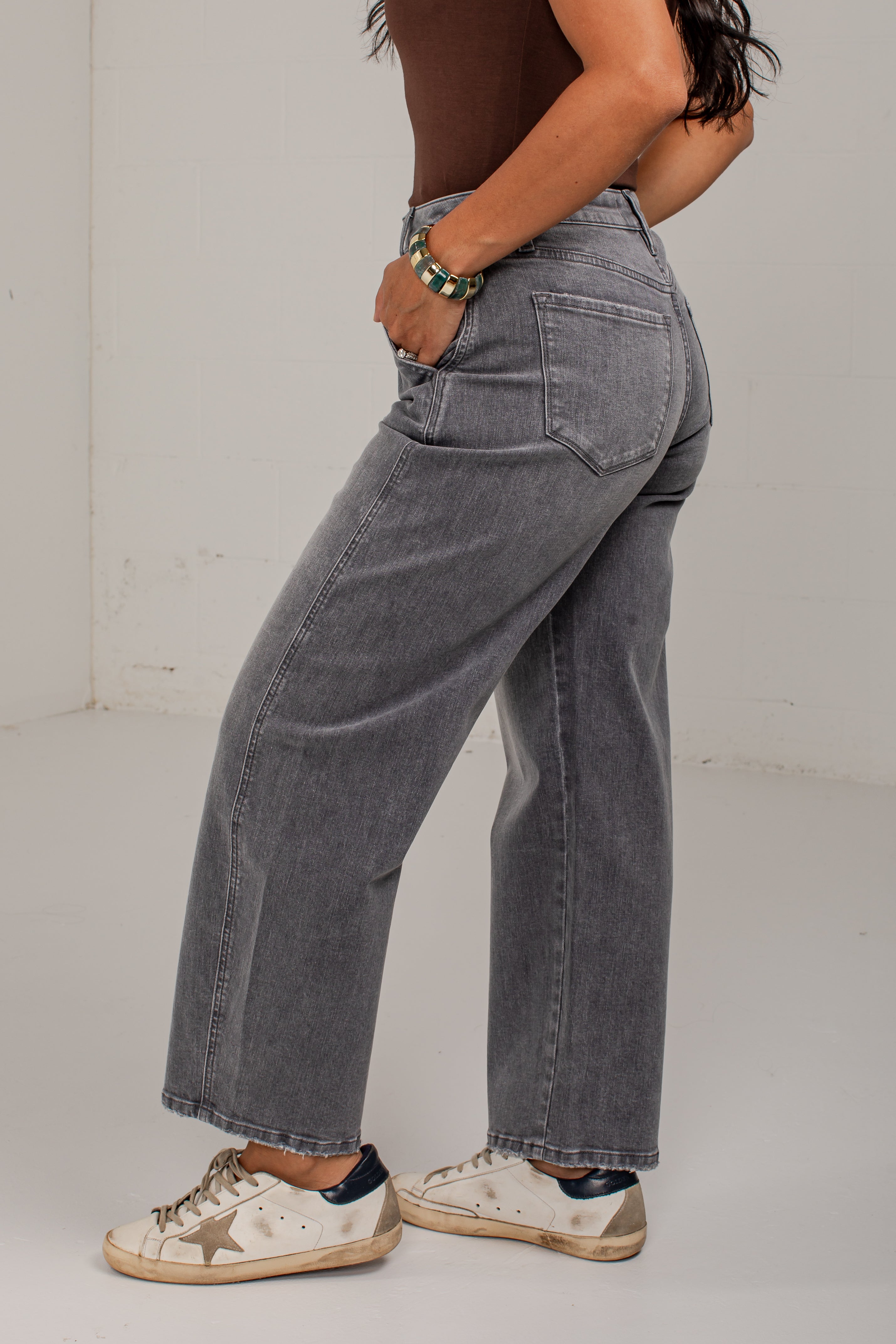 A person wearing gray wide-leg jeans, a brown top, and white sneakers, shown from the side in a studio setting.