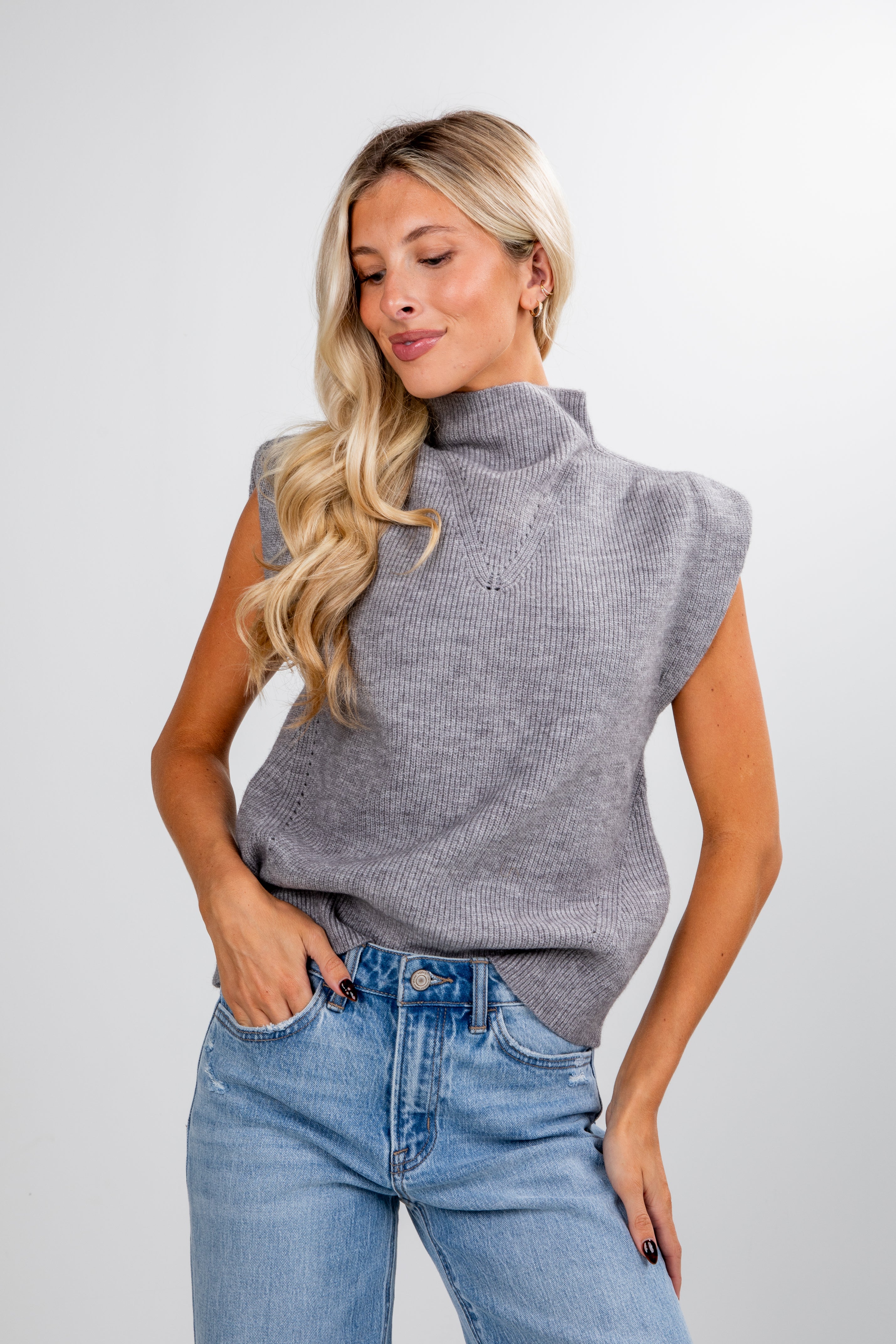 Woman wearing a gray sleeveless sweater and blue jeans against a white background