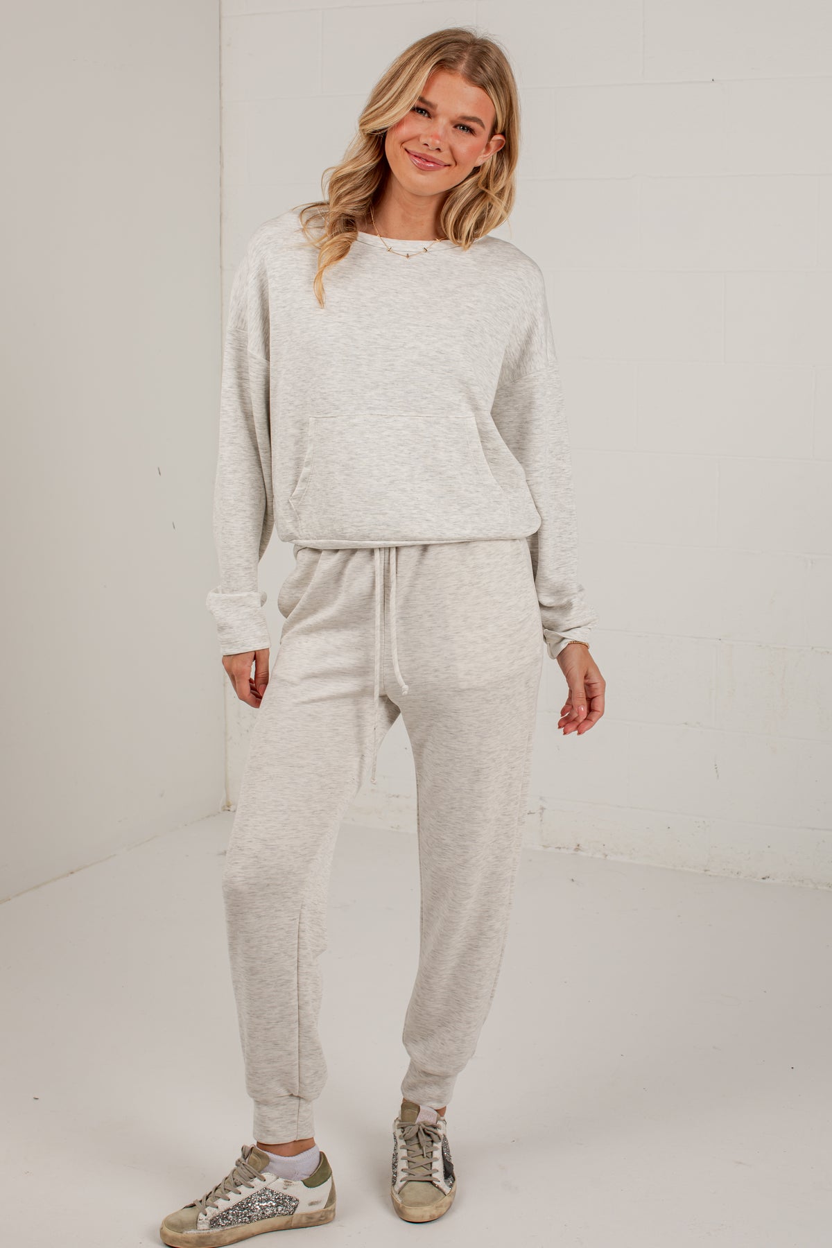 A woman modeling a light heather gray two-piece loungewear set consisting of a long-sleeve sweatshirt and matching jogger pants, paired with distressed sneakers, in a bright studio setting.