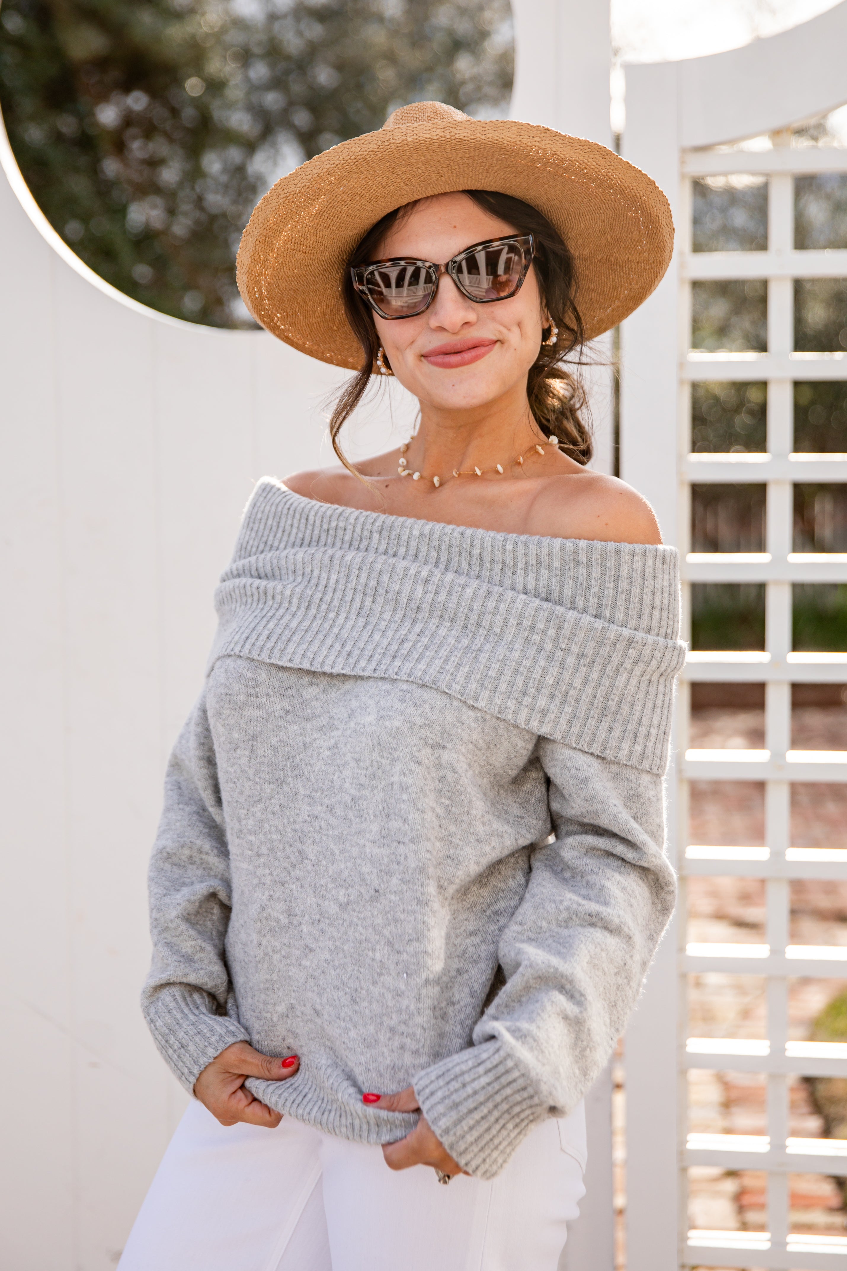 Woman wearing a gray off-shoulder sweater and beige hat with sunglasses, standing against a white lattice background.