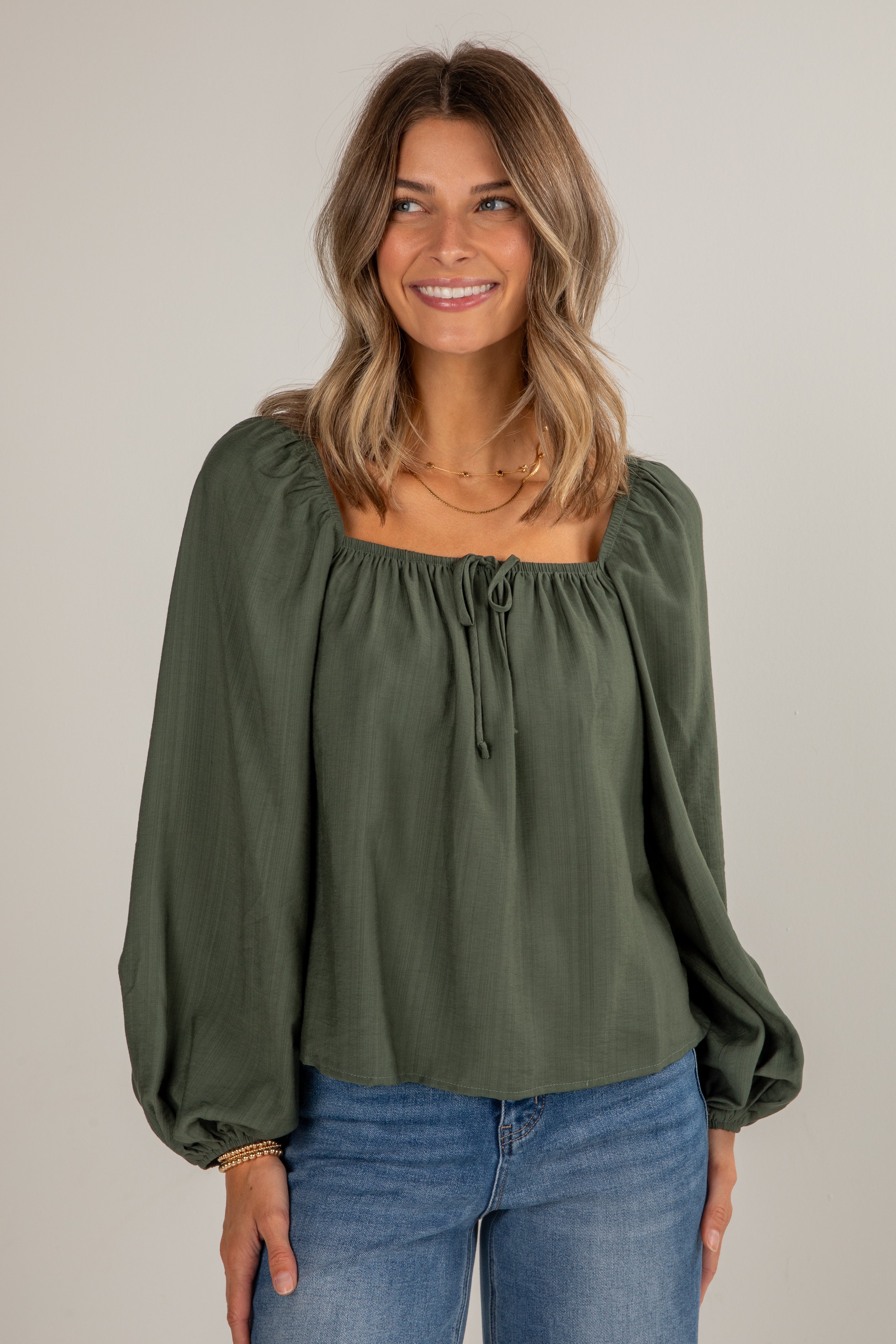 Woman wearing an olive green blouse with long sleeves and blue jeans on a plain background