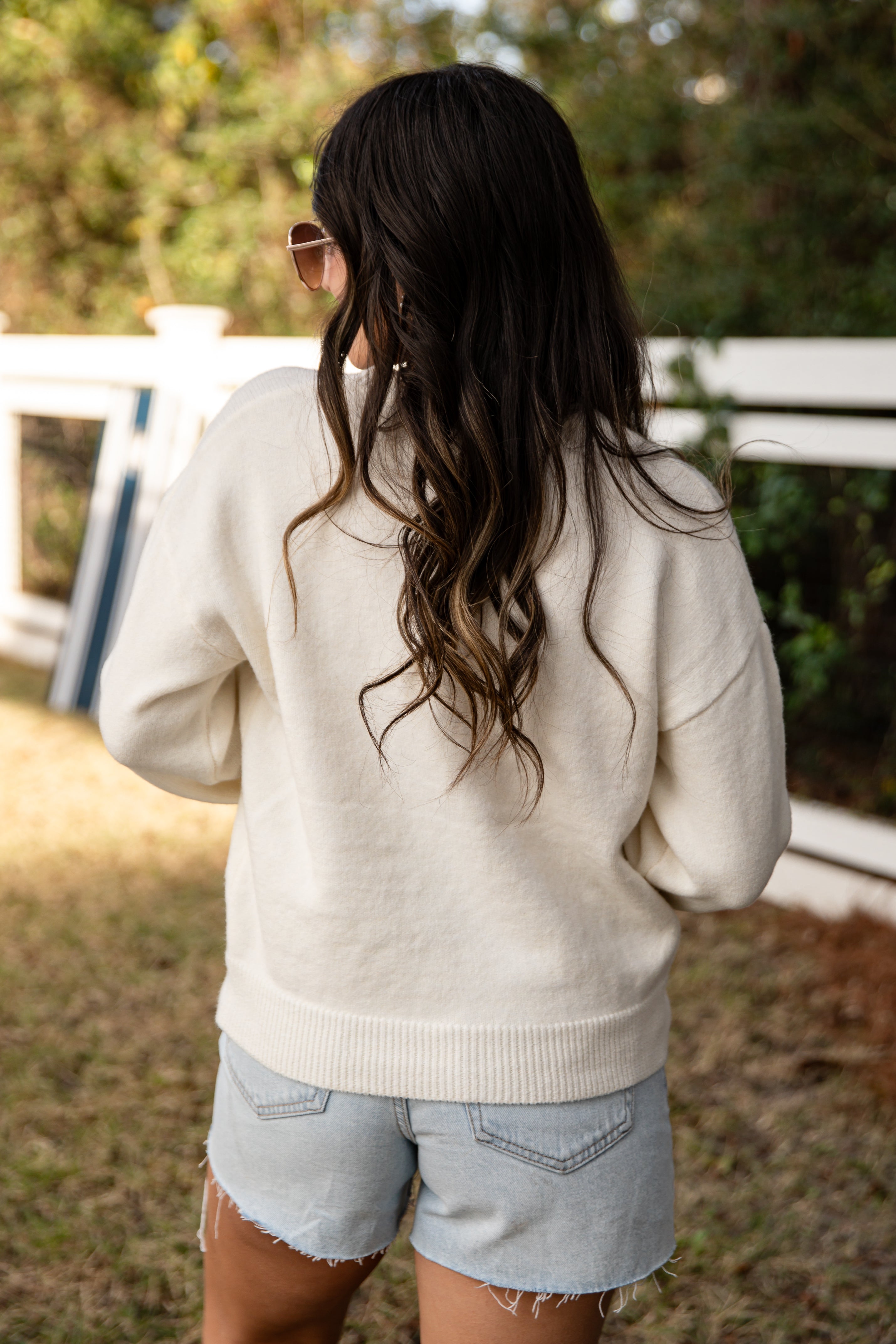 Woman wearing a beige sweater and denim shorts standing outdoors with a blurred background