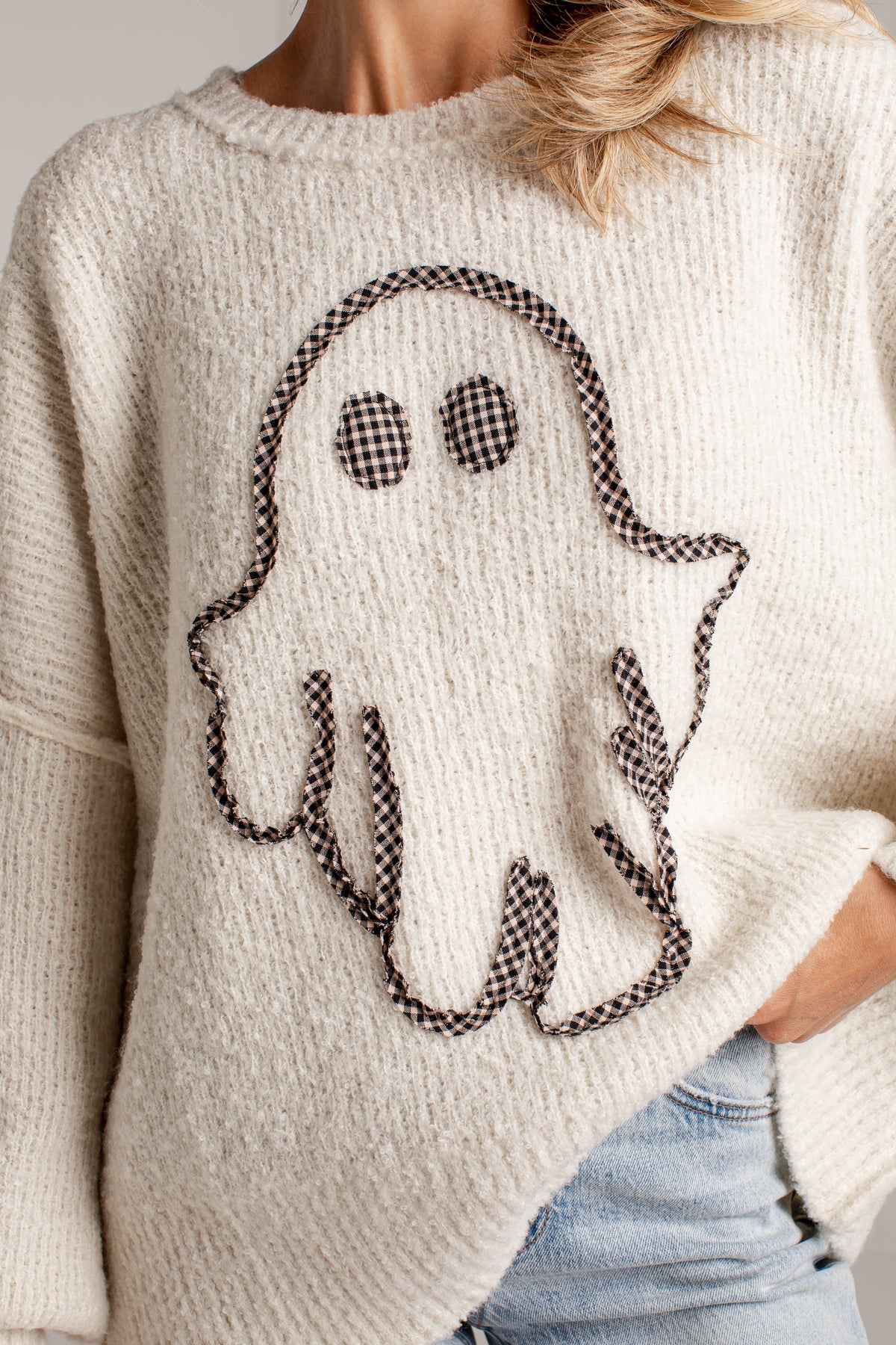 A close-up of a person wearing a cream-colored knit sweater with a black-and-white gingham ghost applique on the front, paired with light blue jeans.