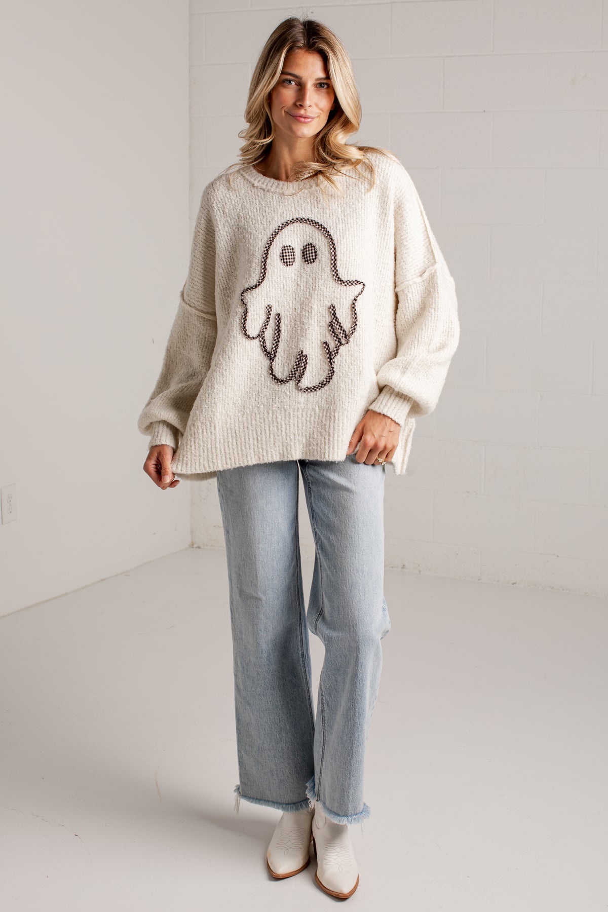A woman modeling a cream-colored knit sweater with a large brown ghost motif on the front, paired with light-wash jeans and white ankle boots, standing against a white brick wall.