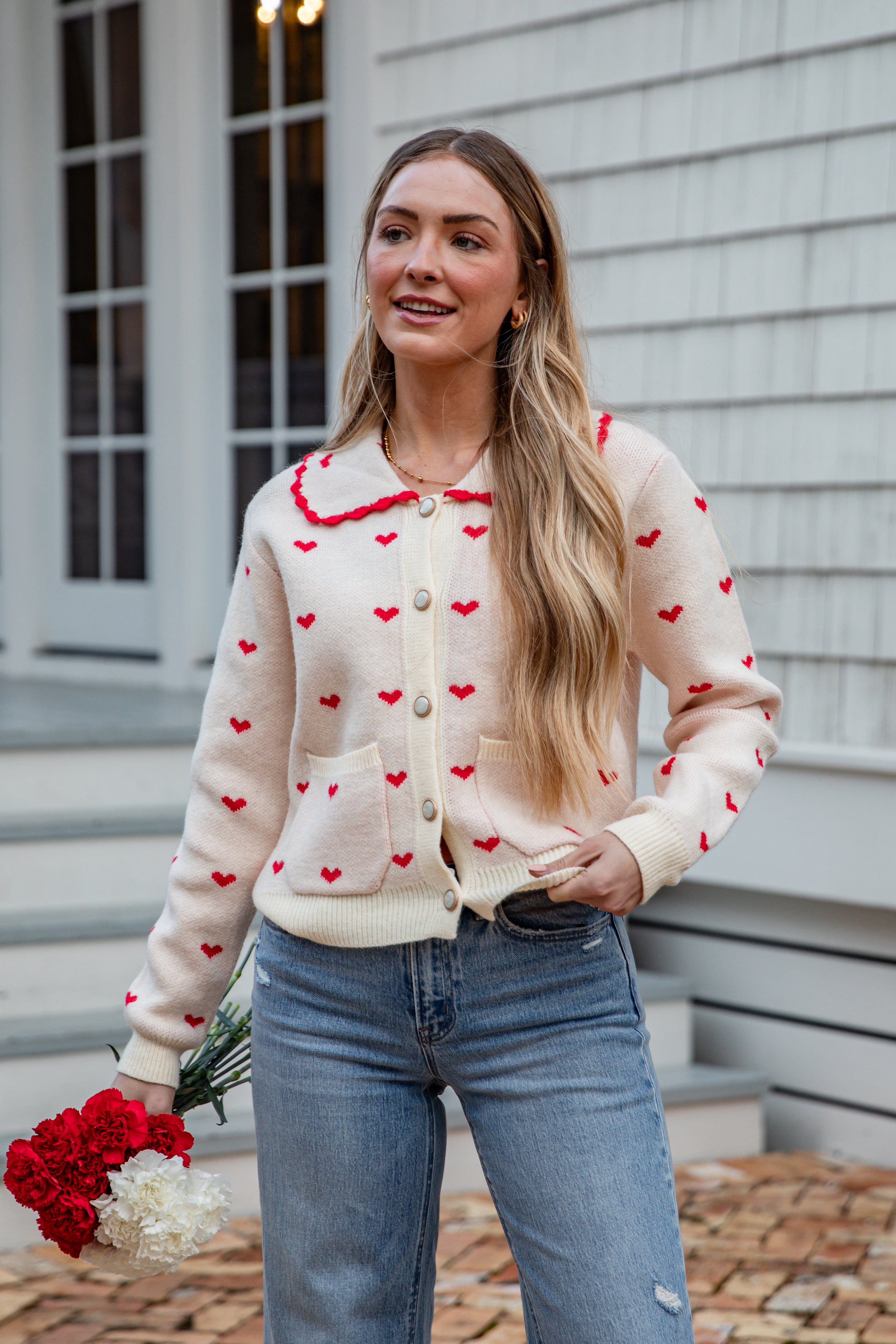 Woman wearing a cream cardigan with red heart patterns and blue jeans, standing outdoors.
